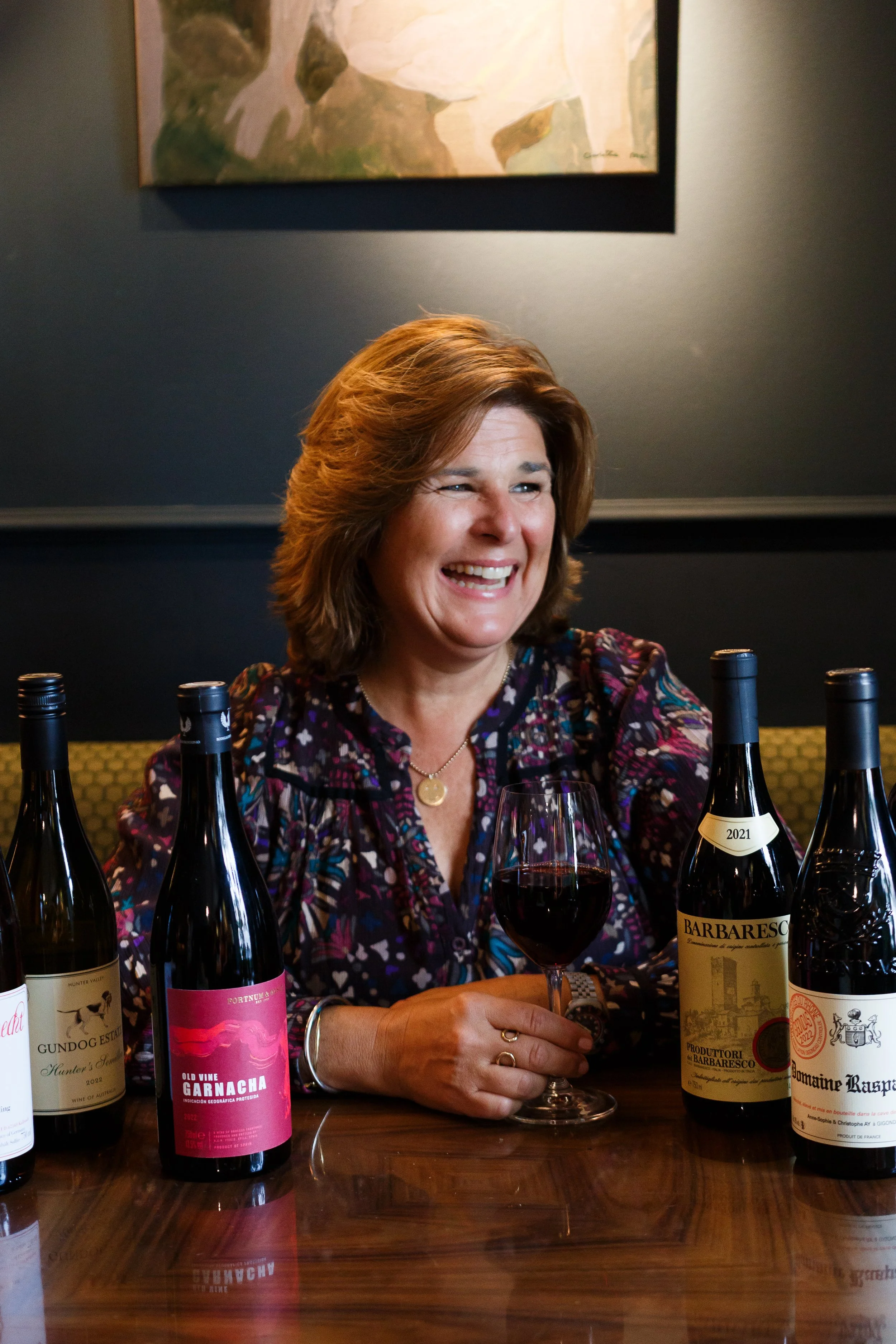A woman with brown hair smiling at a table with several bottles of wine and a glass of red wine. The background has a painting on a dark wall.