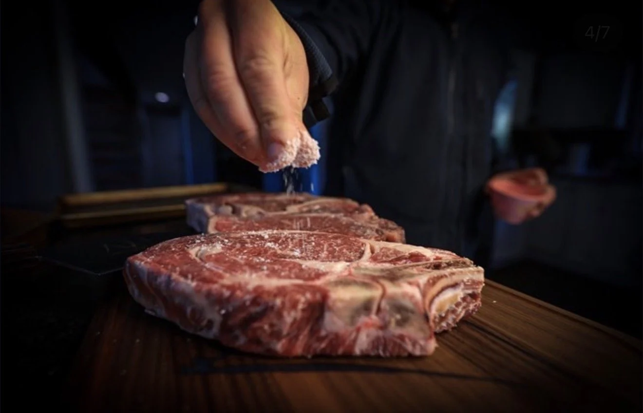 Person seasoning raw beef steaks on a wooden cutting board.