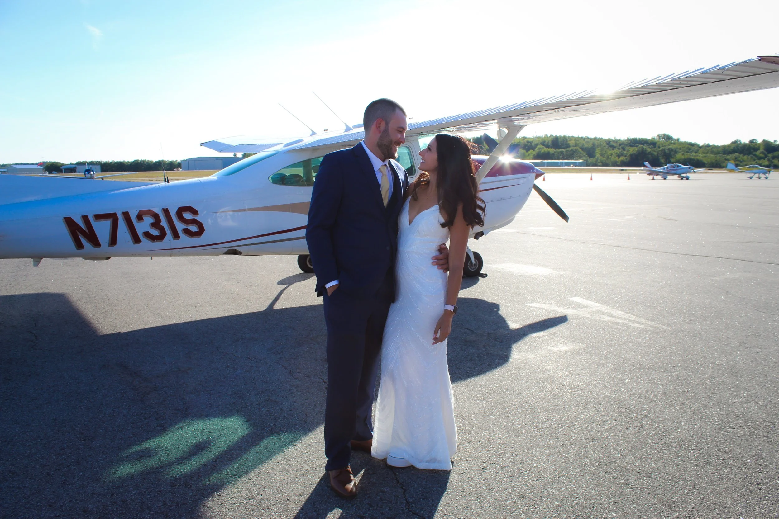 A bride and groom standing close together near a small aircraft on a sunny day, with other planes in the background.