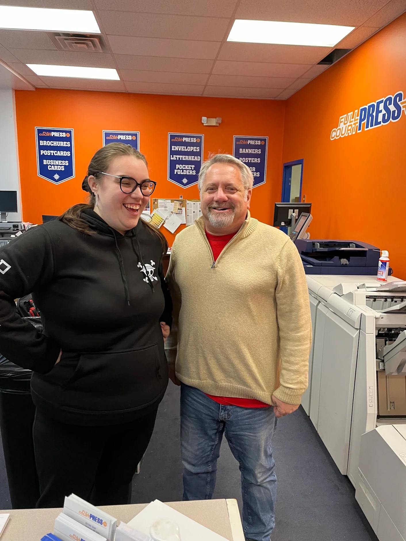 A woman with glasses and a black hoodie and a man in a beige sweater and jeans smiling and standing next to each other in an office with orange walls, printing equipment, and posters about printing services.