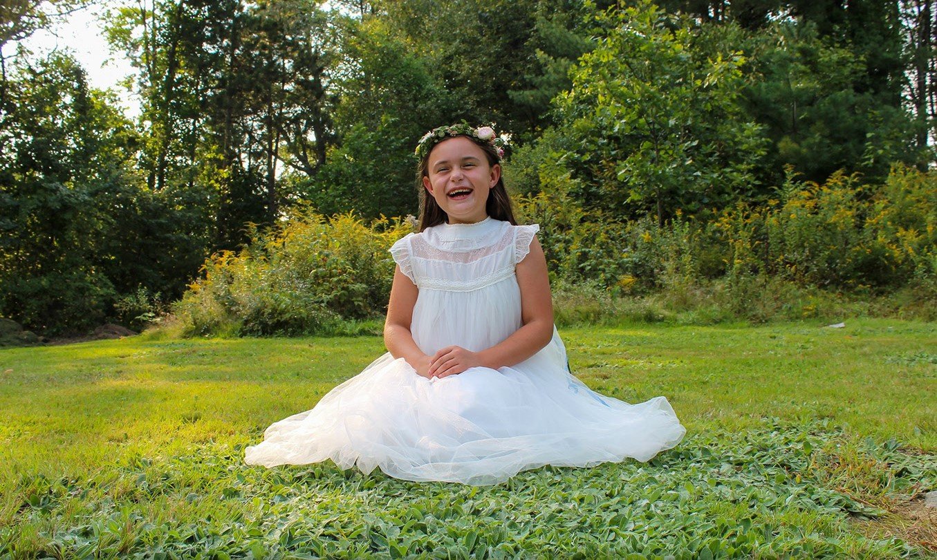A young girl in a white dress sitting on grass outdoors, smiling, wearing a floral crown, with trees in the background.