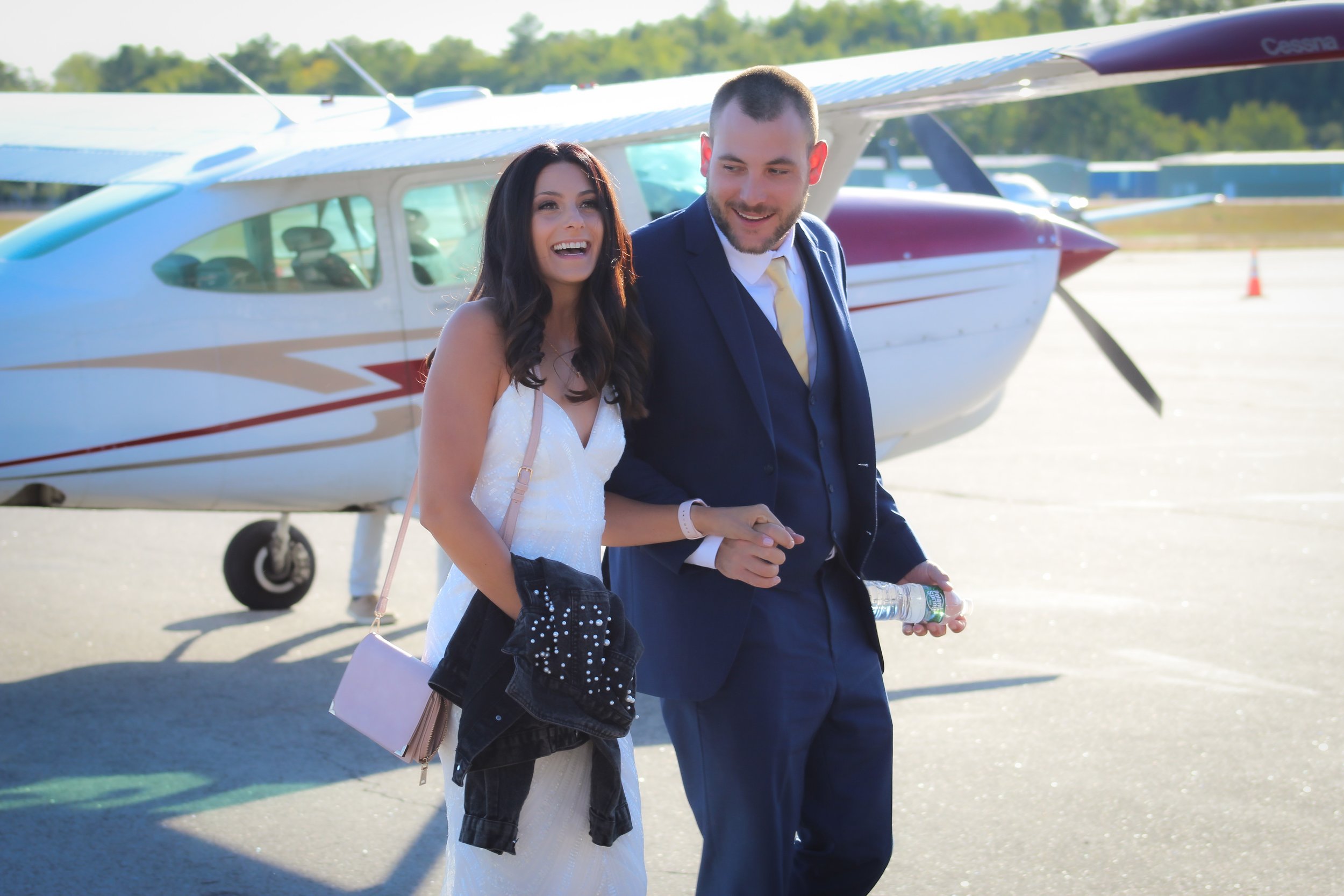 A smiling woman in a white dress and a man in a dark suit walking on an airport tarmac with a small aircraft in the background.