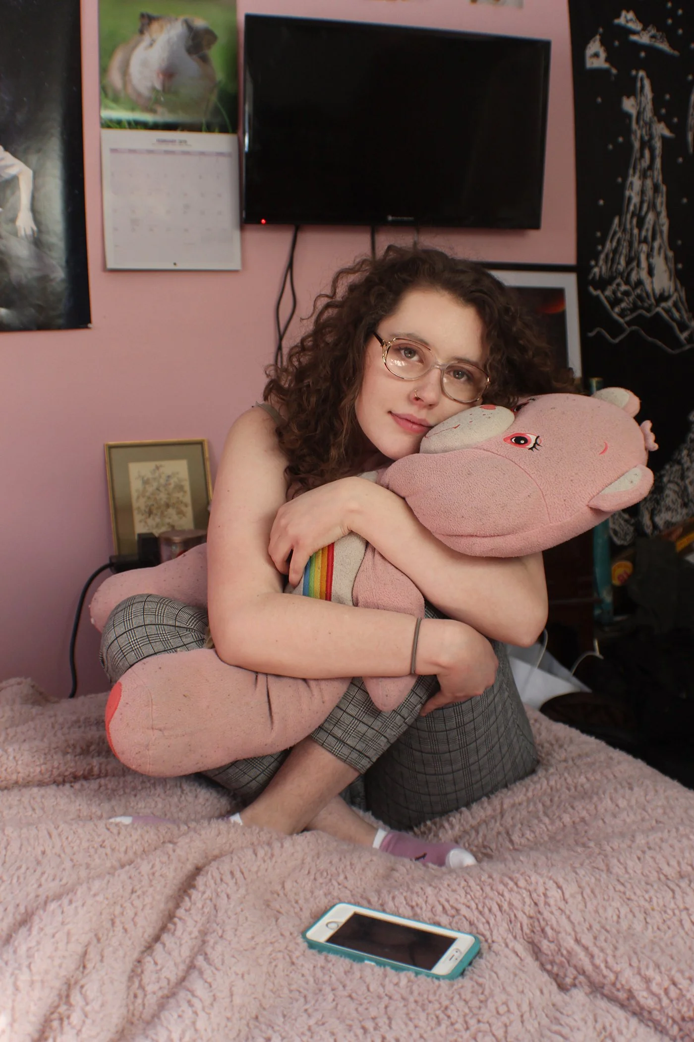 A young woman with curly brown hair, glasses, and a nose ring, hugging a large pink plush toy with a rainbow stripe on its side in a room with pink walls. Behind her are posters, a black television, and a framed picture. There is a smartphone on a pi