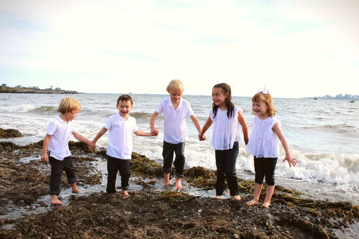 Five children in white tops and dark pants holding hands and playing on a rocky beach near the ocean, with waves in the background.