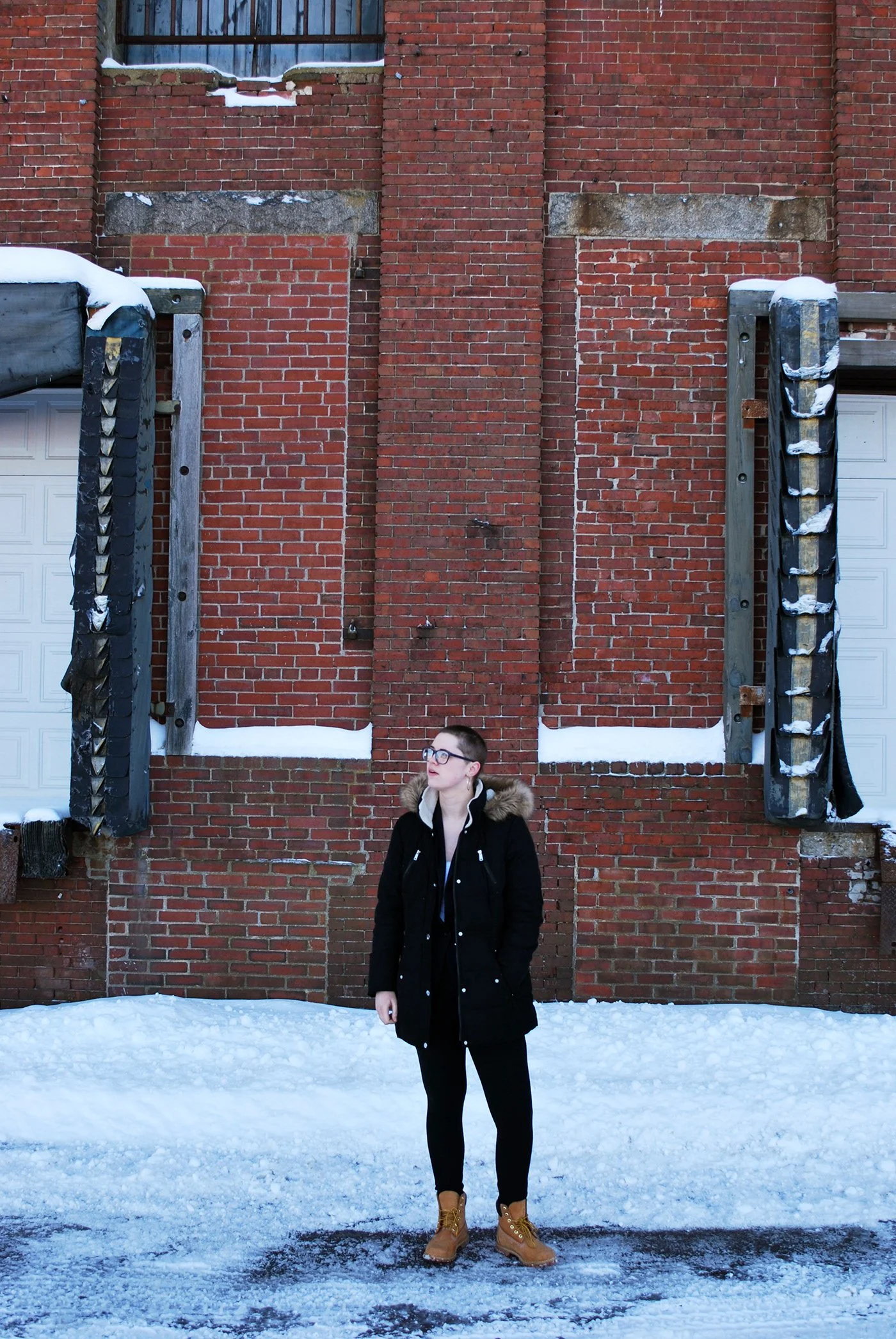 A person wearing a black coat, tan boots, and glasses standing in front of a brick wall with snow on the ground and on the wall structures.