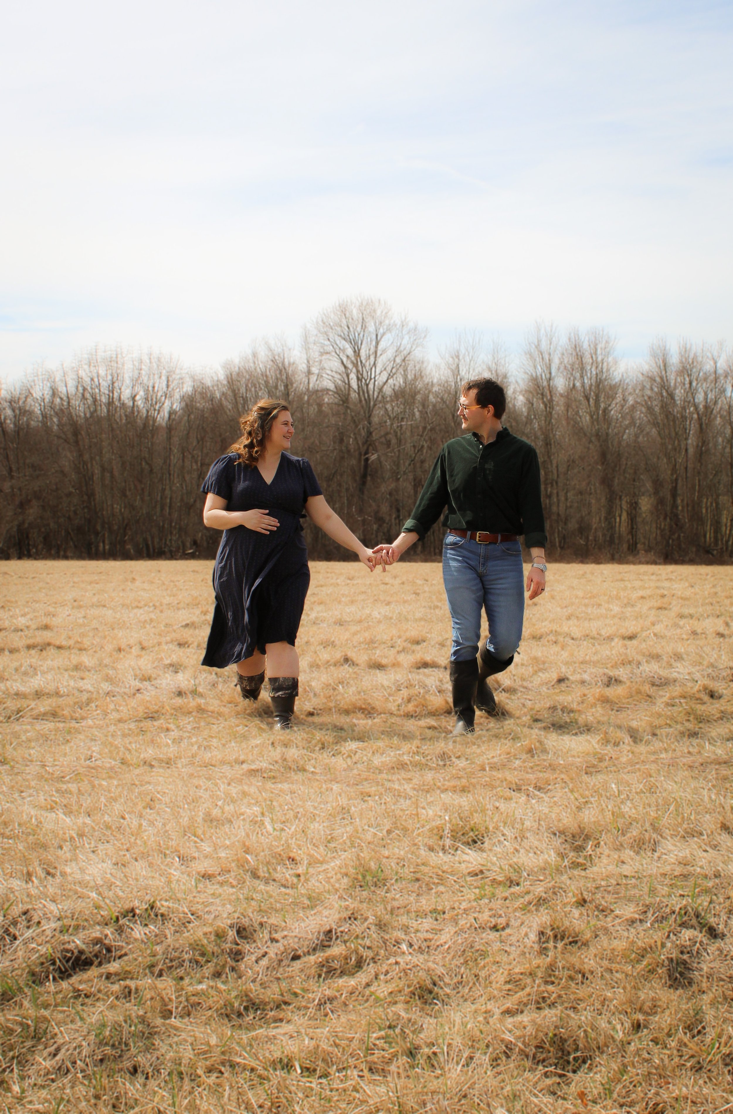 A man and woman holding hands walking through a dry grassy field, with leafless trees in the background under a partly cloudy sky.