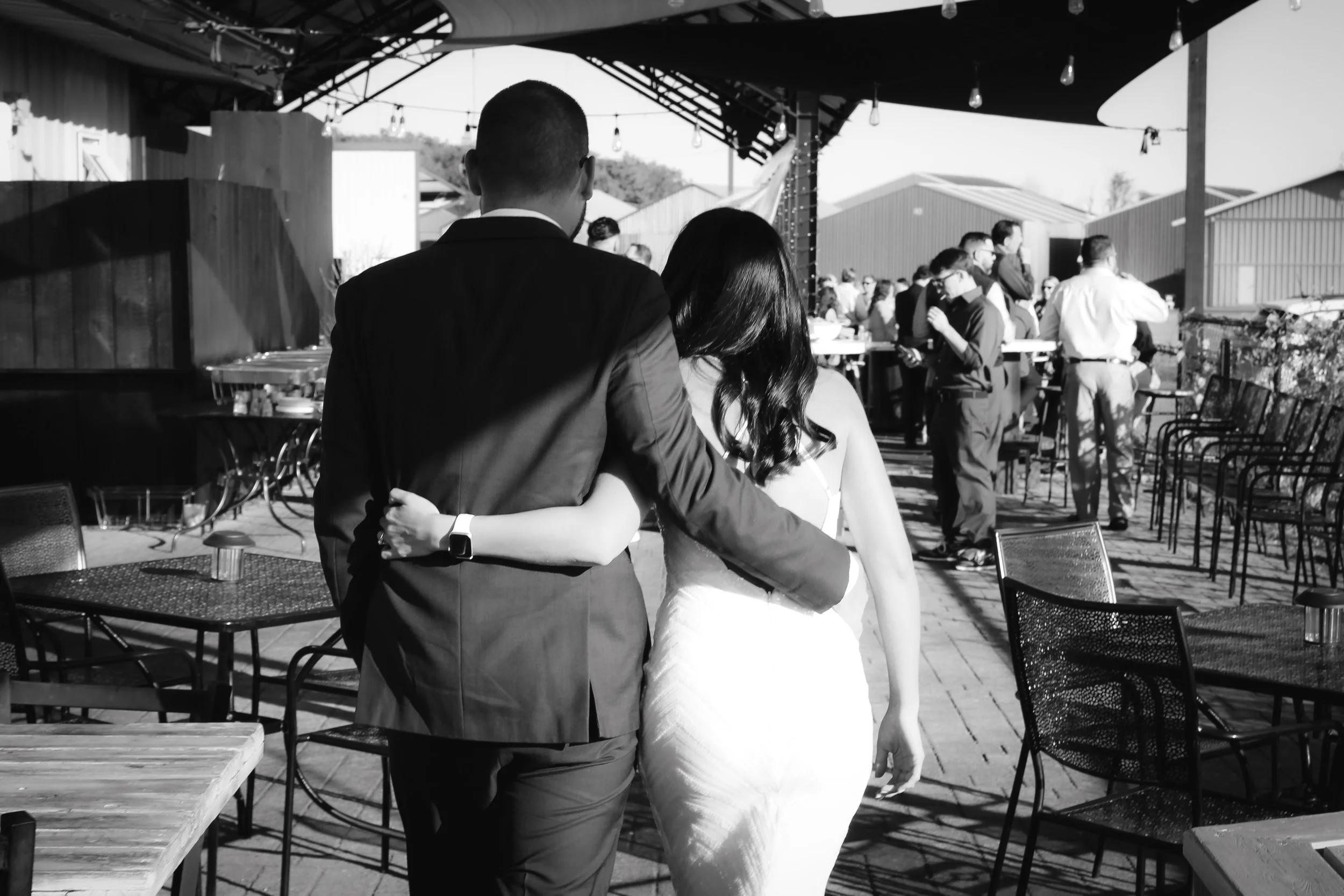A black and white photo of a couple with the man in a suit and the woman in a wedding dress, embracing at an outdoor wedding reception.