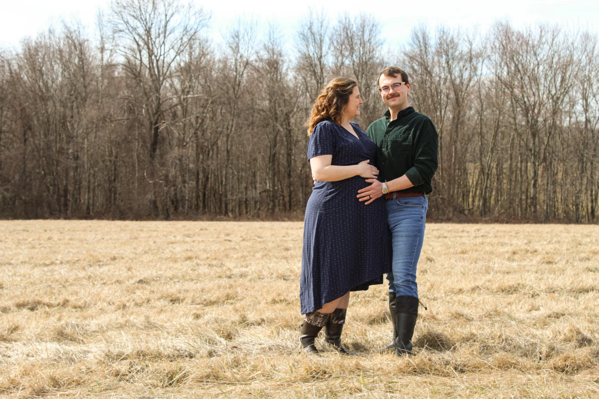 A pregnant woman and a man stand together in a field, embracing and smiling. The woman wears a blue dress and cowboy boots; the man wears a dark shirt, jeans, and boots. Bare trees are in the background.