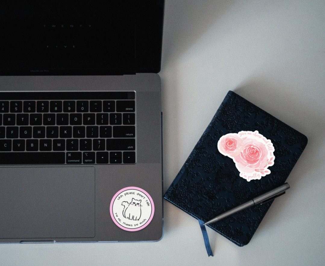 A laptop on a white desk with a round sticker of a cat and text, a closed navy blue floral notebook with a pink rose sticker, and a silver pen.