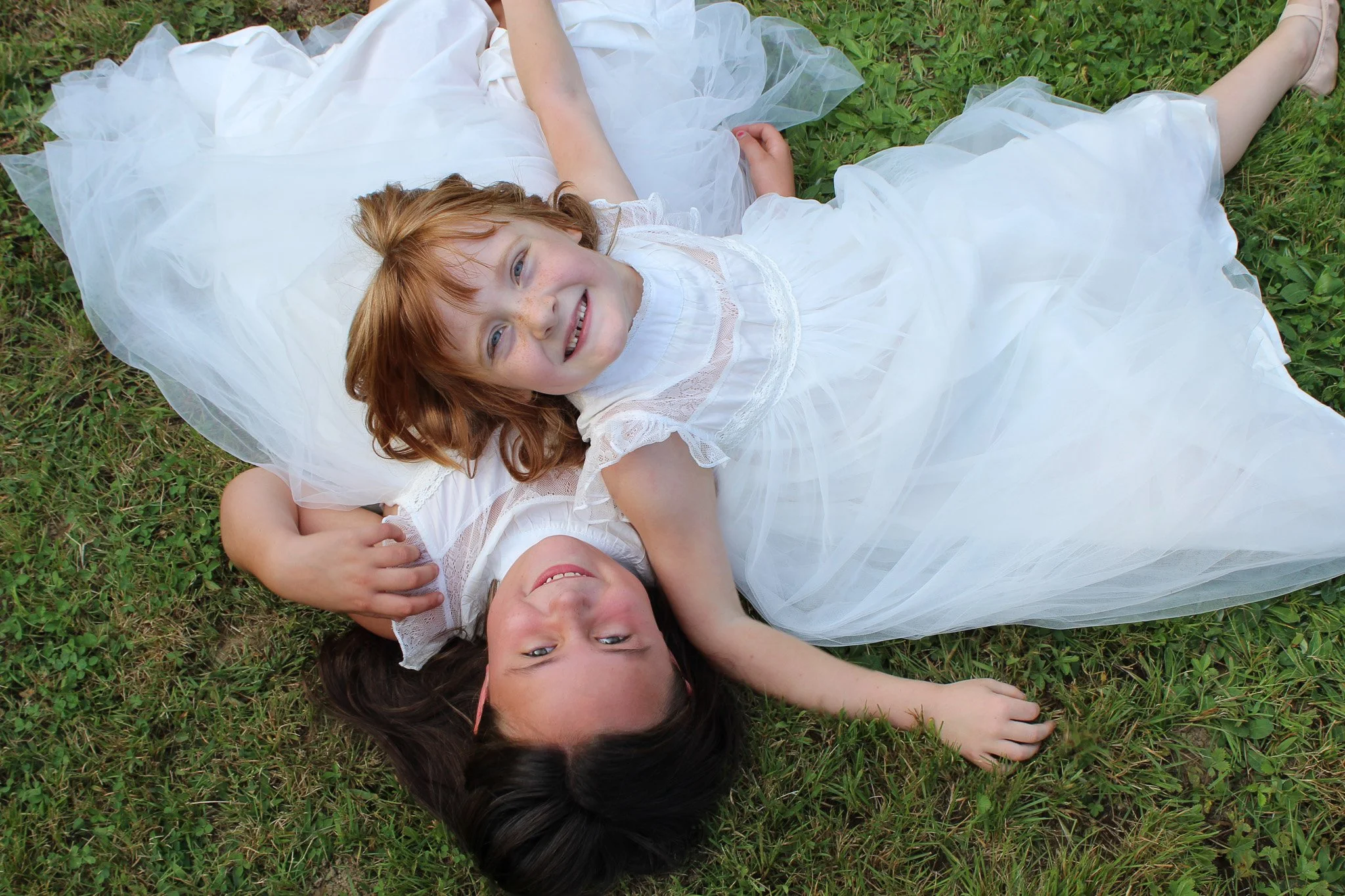 Three young girls lying on grass, one in a white dress, smiling and looking up at the camera.