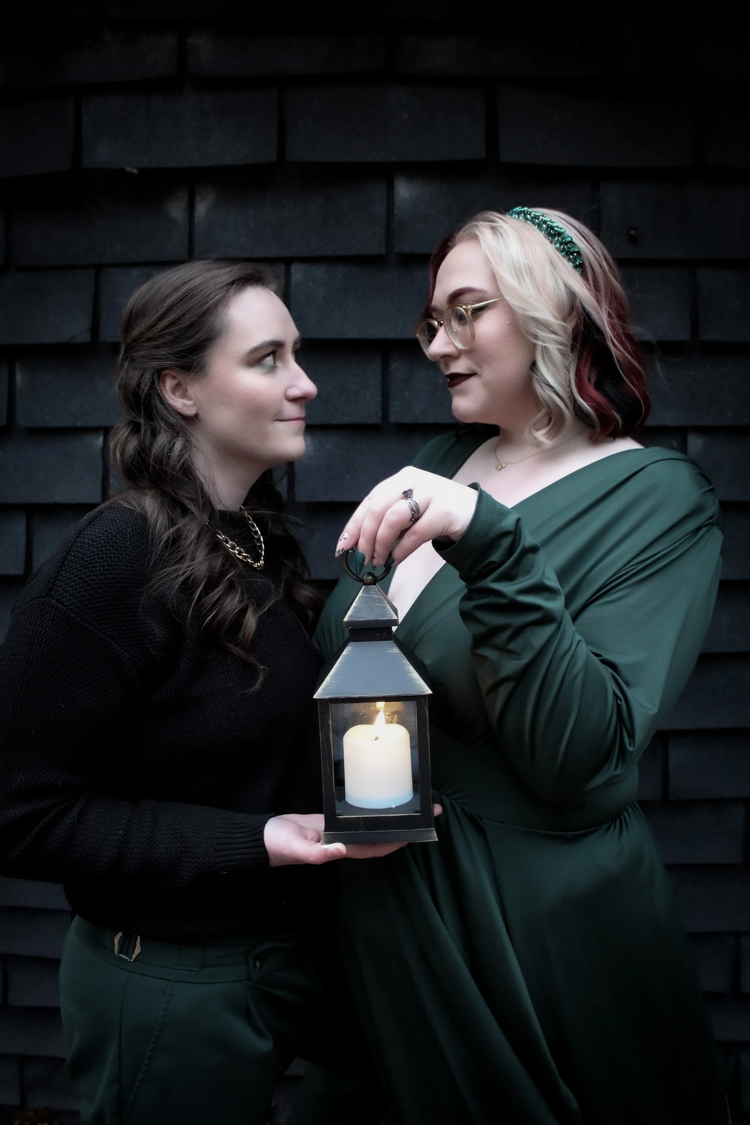 Two women standing close to each other, holding a lantern with a lit candle inside, against a dark brick wall background.