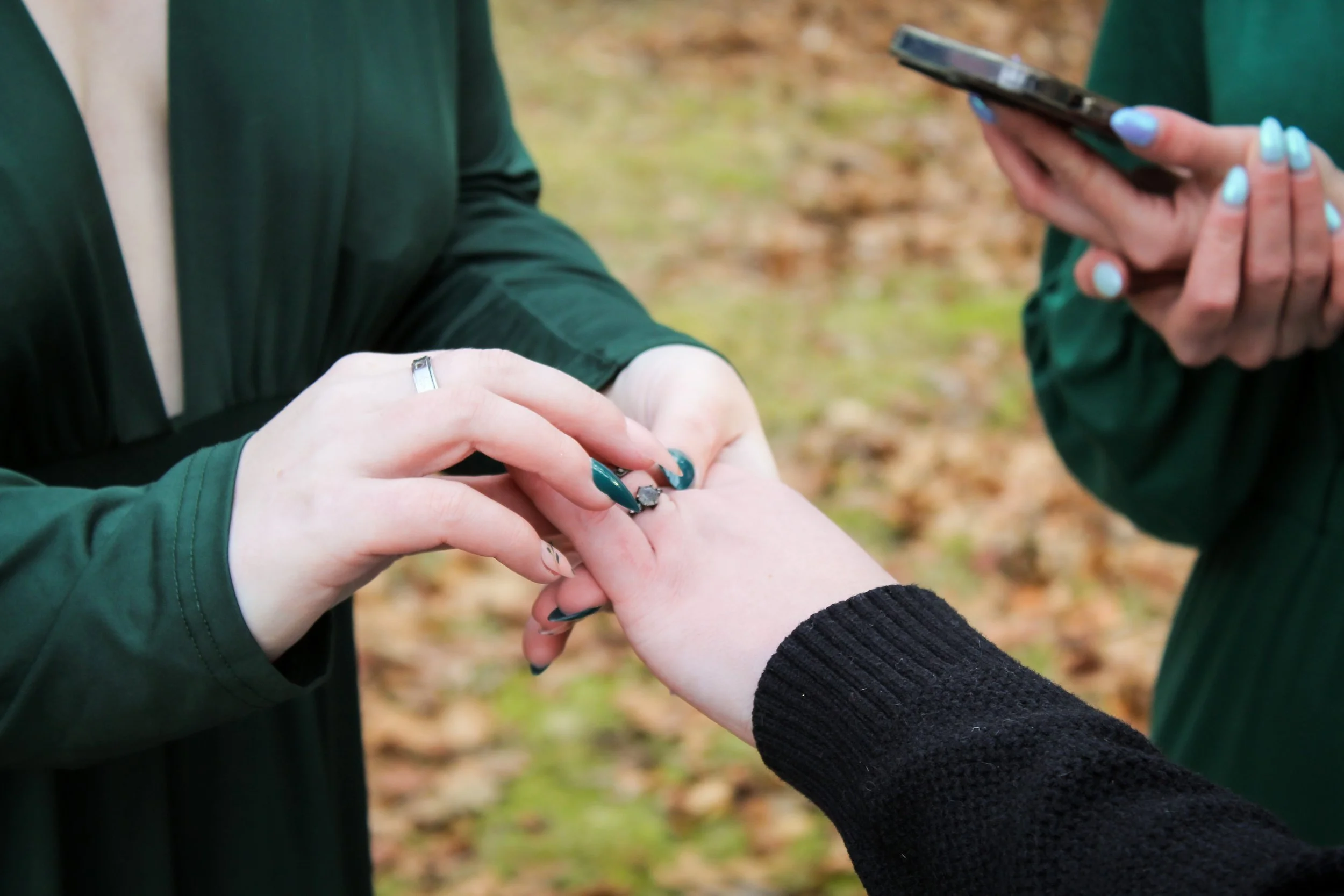 Two women exchanging rings outdoors, with one woman placing a ring on the other's finger and another woman holding a phone nearby.