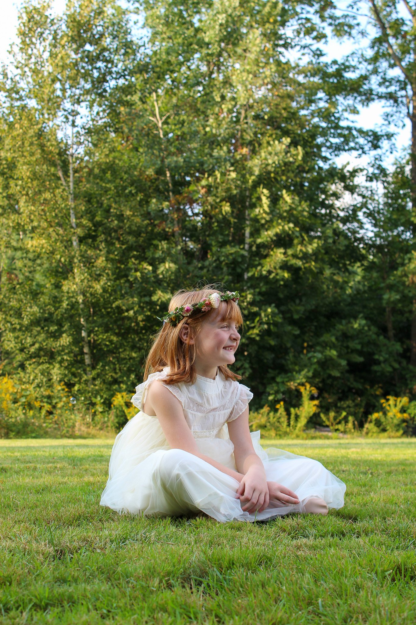 A young girl with red hair, wearing a white dress and a floral crown, sitting on the grass in a park with green trees in the background.