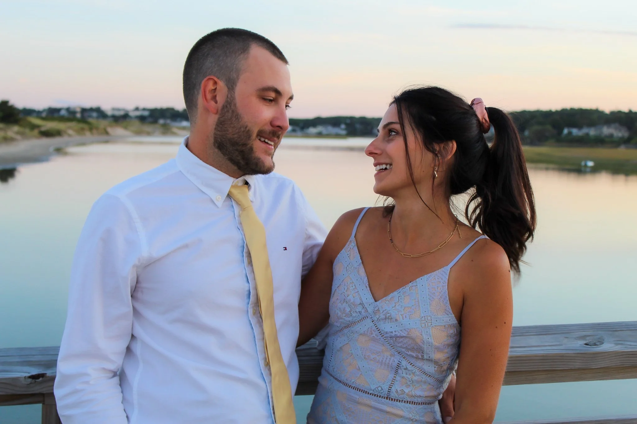 A man and woman sharing a joyful moment by a body of water during sunset. The man is wearing a white shirt and yellow tie, and the woman is in a white lace dress with earrings and a necklace.