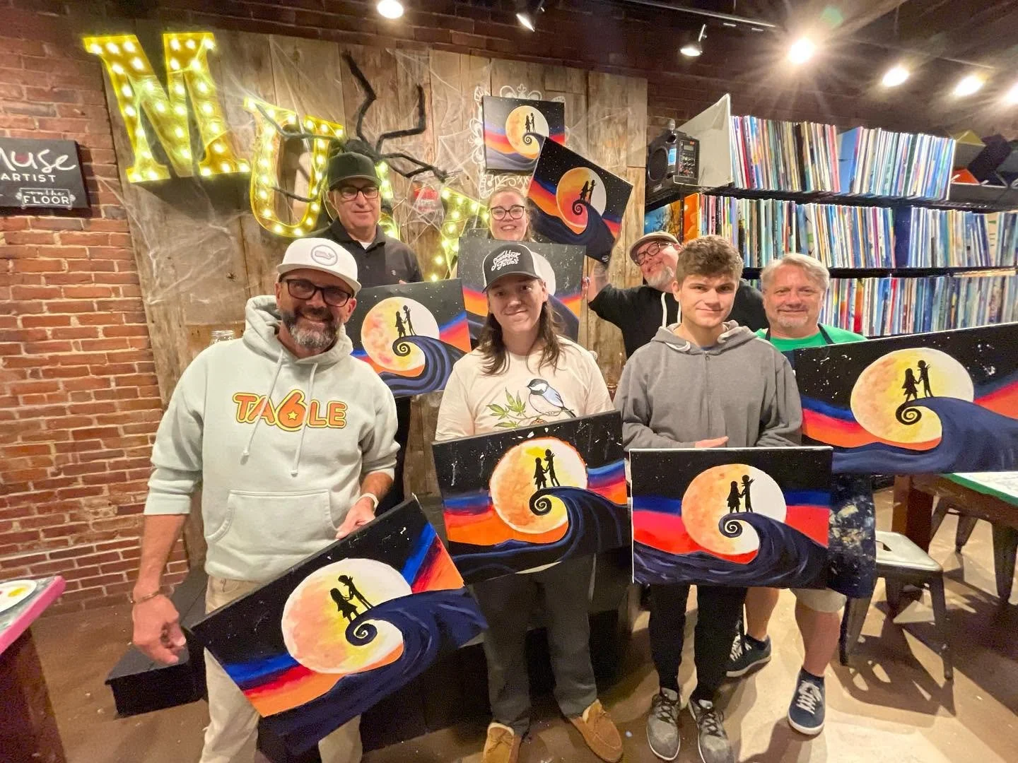 Group of people at an art event showing their completed paintings of a whimsical night scene with a large orange moon, a silhouetted couple holding hands, and swirling clouds in vibrant colors.