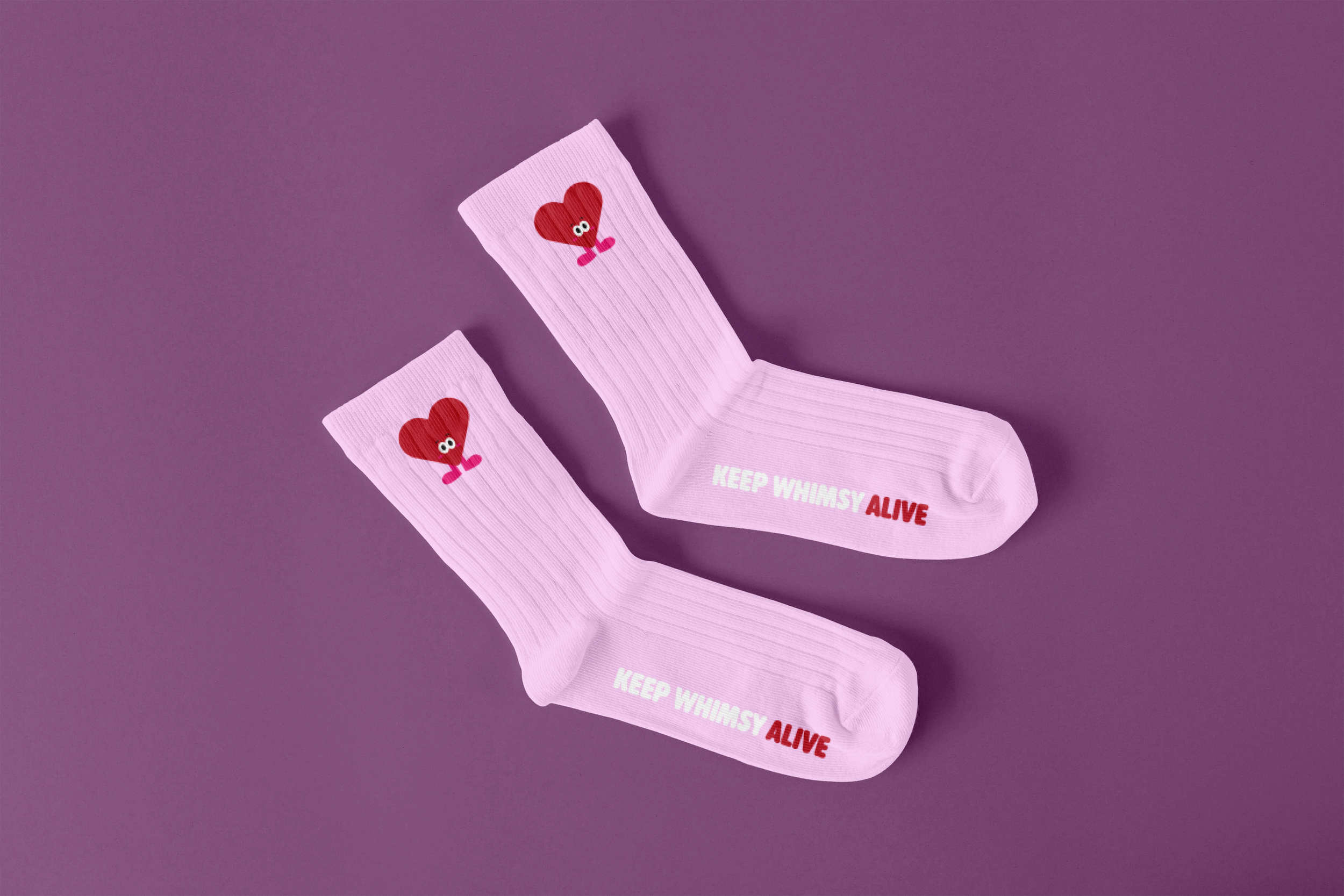 Pink socks with a red heart character with eyes and legs, laid on a purple surface. The socks have the phrase 'KEEP WHIMSY ALIVE' printed on them.