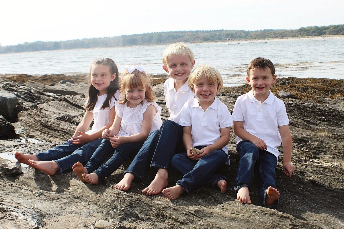 Six children sitting on a rocky shore by the water, smiling at the camera, wearing white tops and jeans.