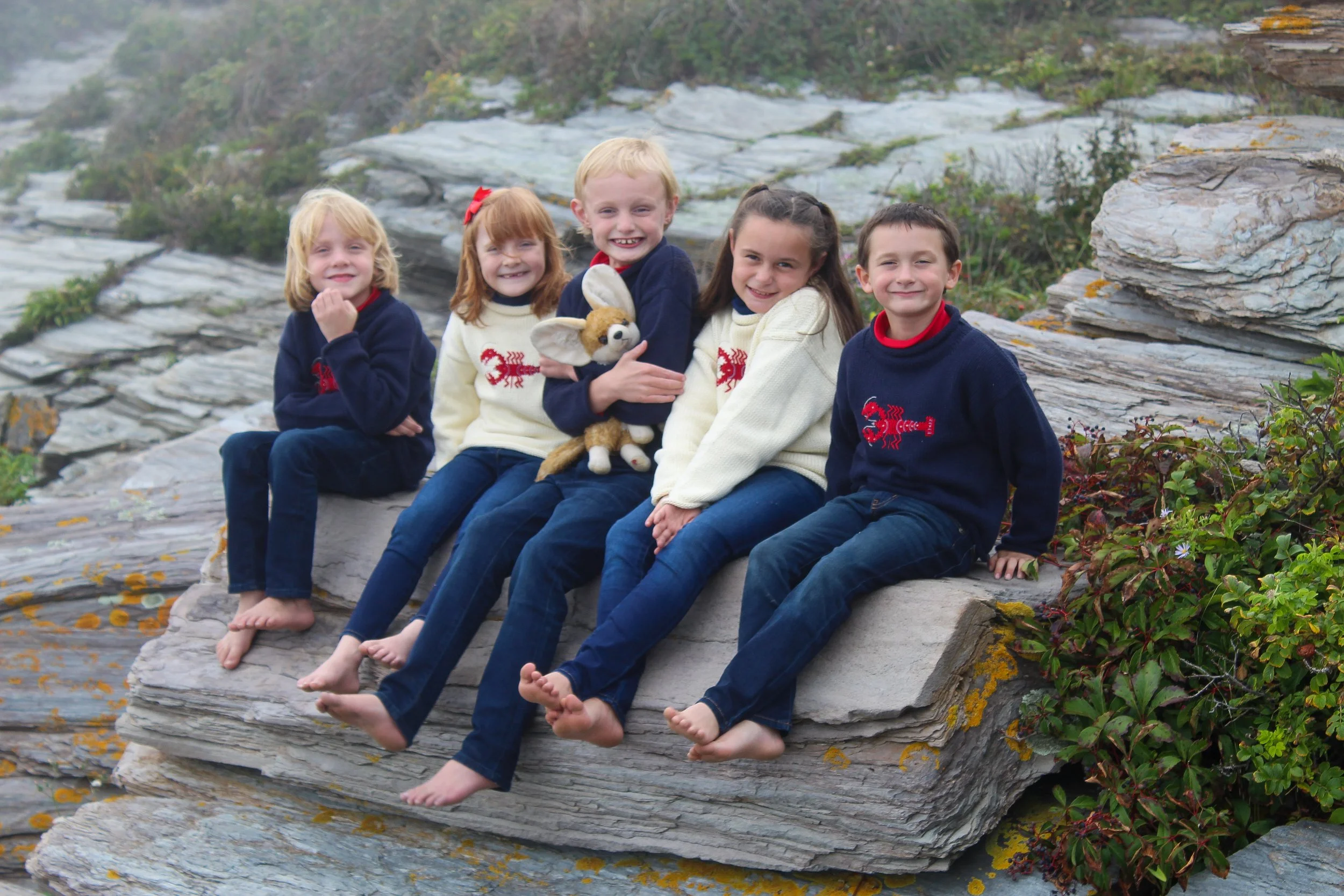 Six children sitting on rocks by the water, holding a stuffed bunny, smiling, with greenery around.