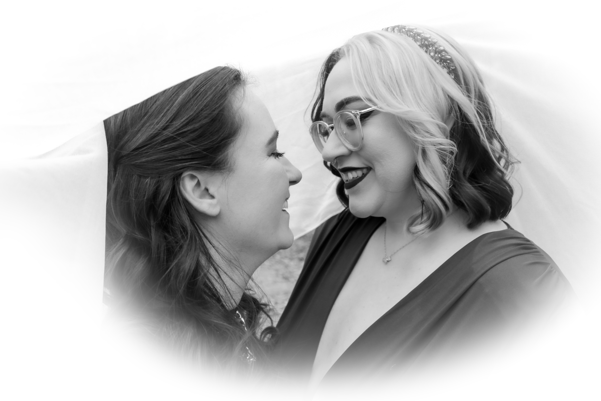 Two women face each other closely, smiling, with their foreheads almost touching, outdoors, with a blurred background.