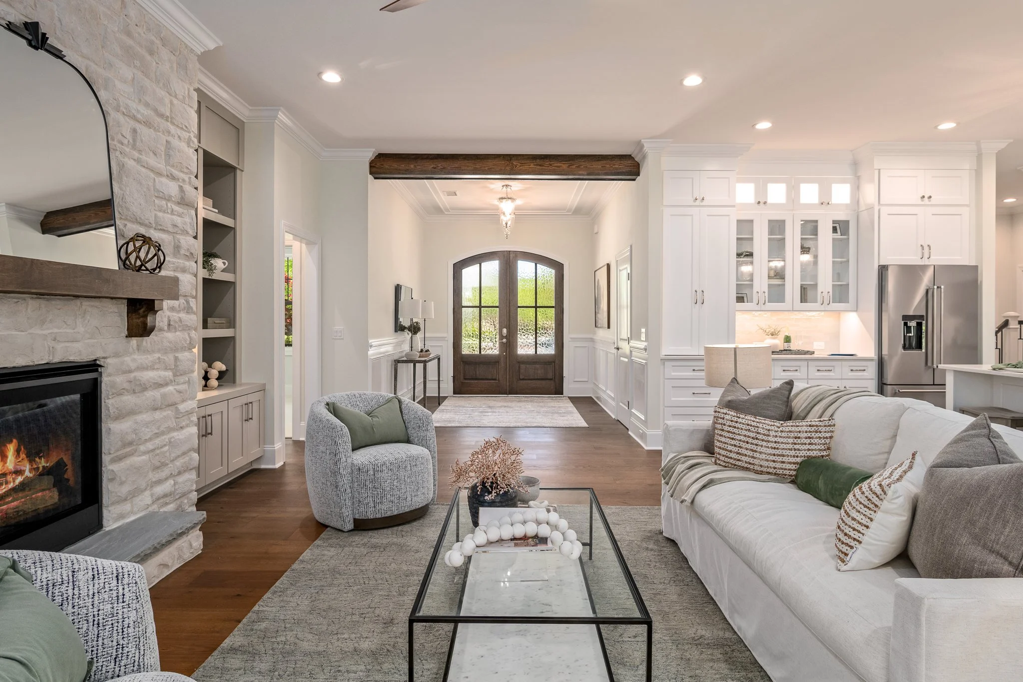 Living room with white walls and cabinetry, wooden flooring, a fireplace on the left, modern furniture including a white sofa and patterned armchair, and a large front door with glass panes.