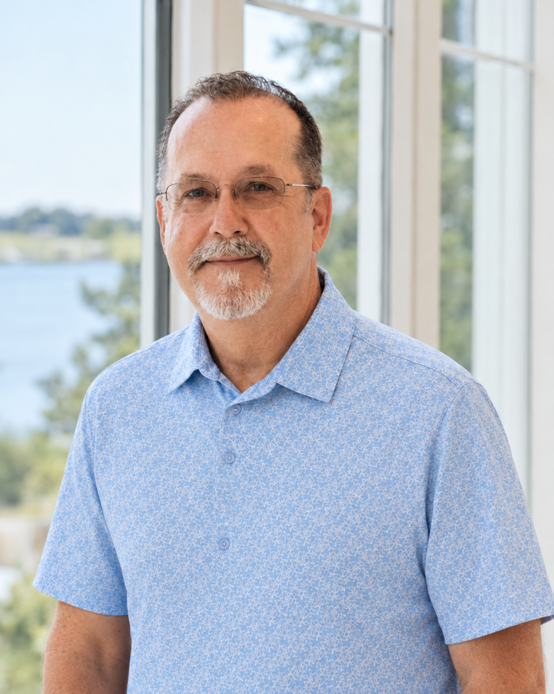 A middle-aged man with glasses and a white beard stands in front of a large window with a view of trees and water, wearing a light blue patterned short-sleeve shirt.