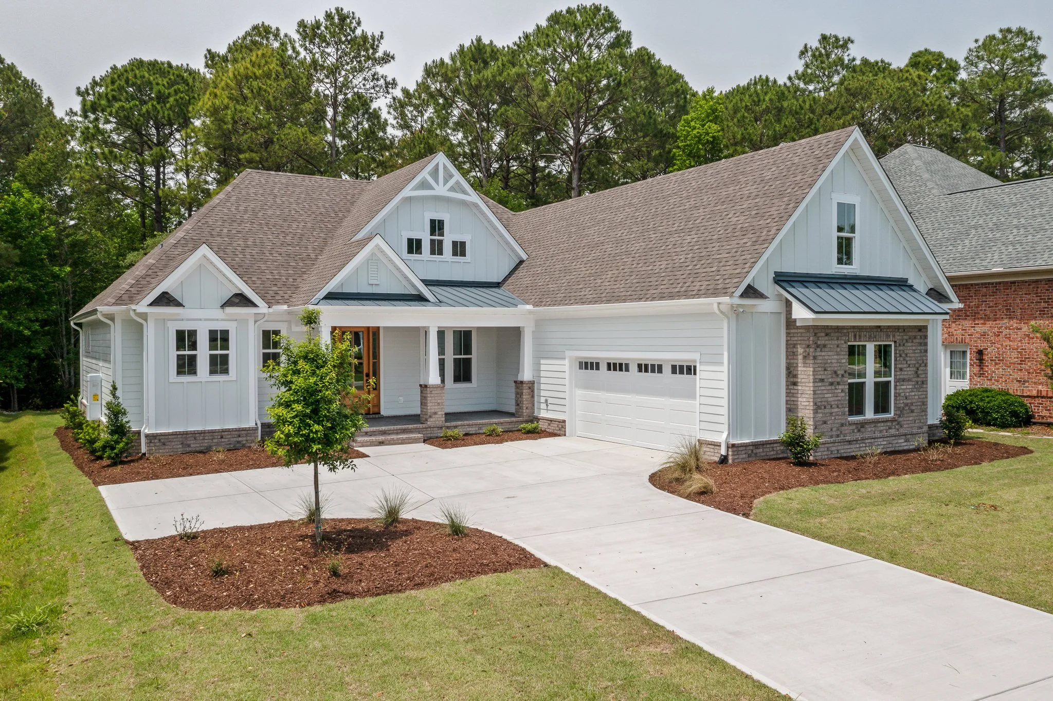 Front view of a two-story house with white siding, brick accents, a gabled roof with gray shingles, and a two-car garage, surrounded by green lawns and trees.