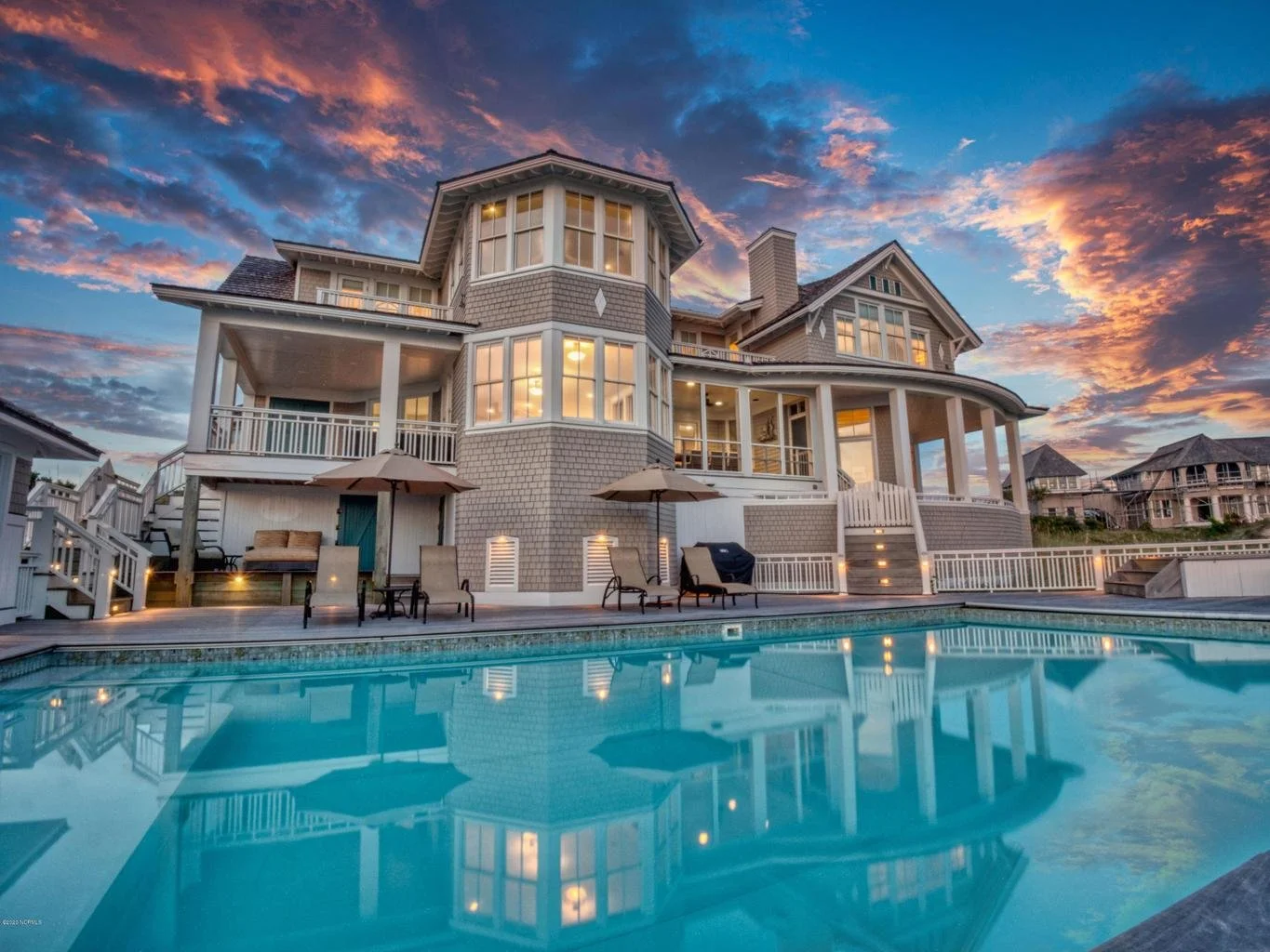 A large, multi-story house with numerous windows, a porch, and a telescope on the balcony, situated behind a swimming pool with lounge chairs and umbrellas, under a colorful sunset sky.