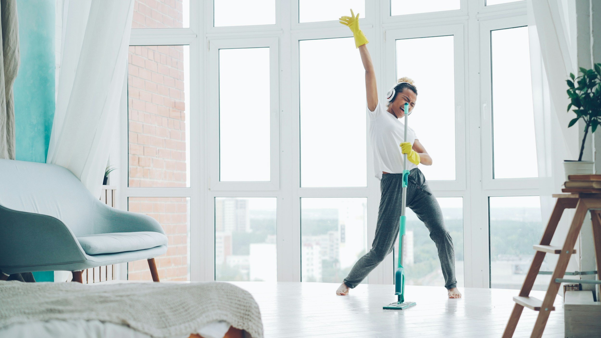Woman cleaning a large window with a mop, wearing headphones, yellow cleaning gloves, and casual clothing, in a bright, modern living room.