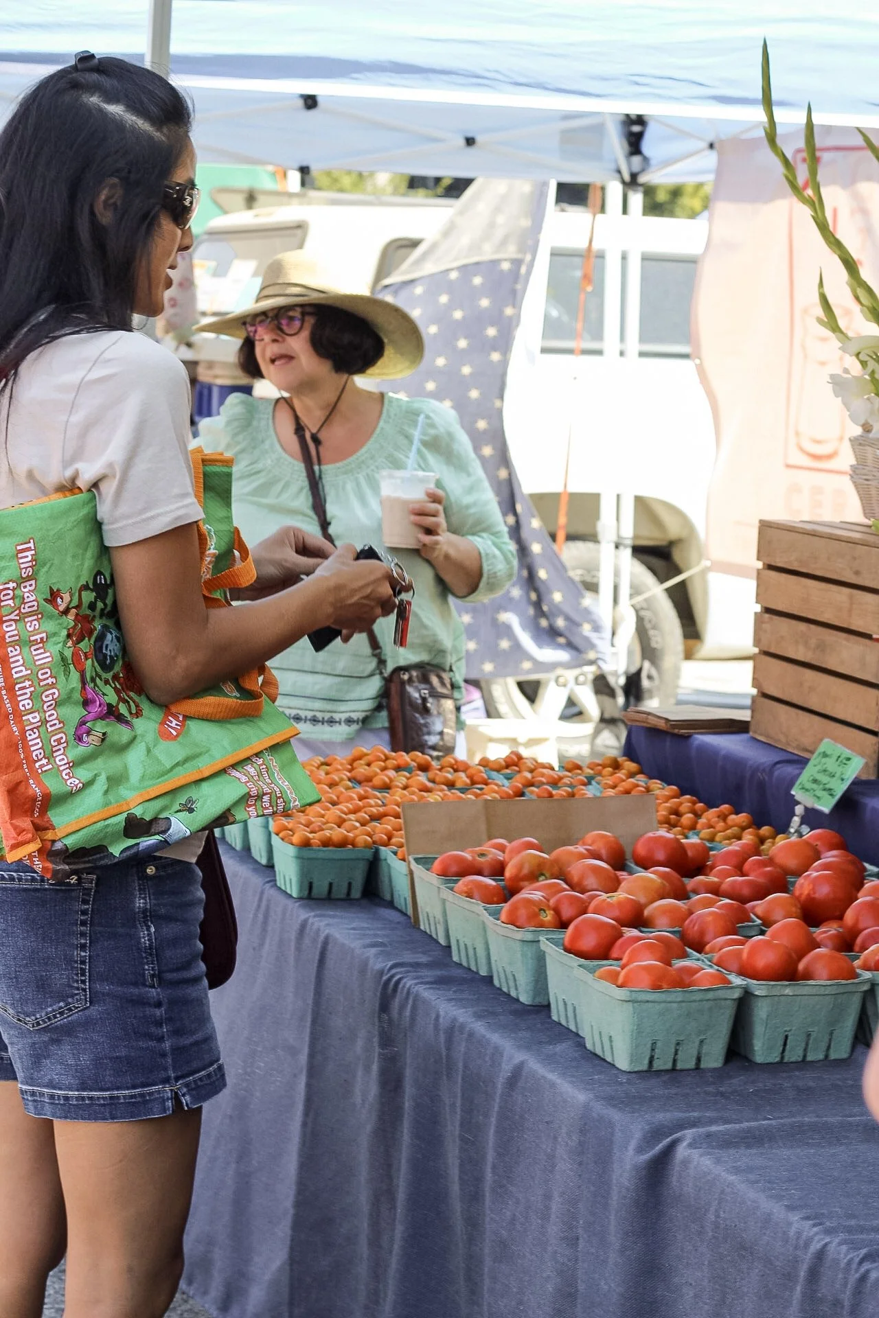 A woman with dark hair, wearing sunglasses, a white shirt, and denim shorts, at an outdoor farmers market stall with baskets of tomatoes and cherry tomatoes.