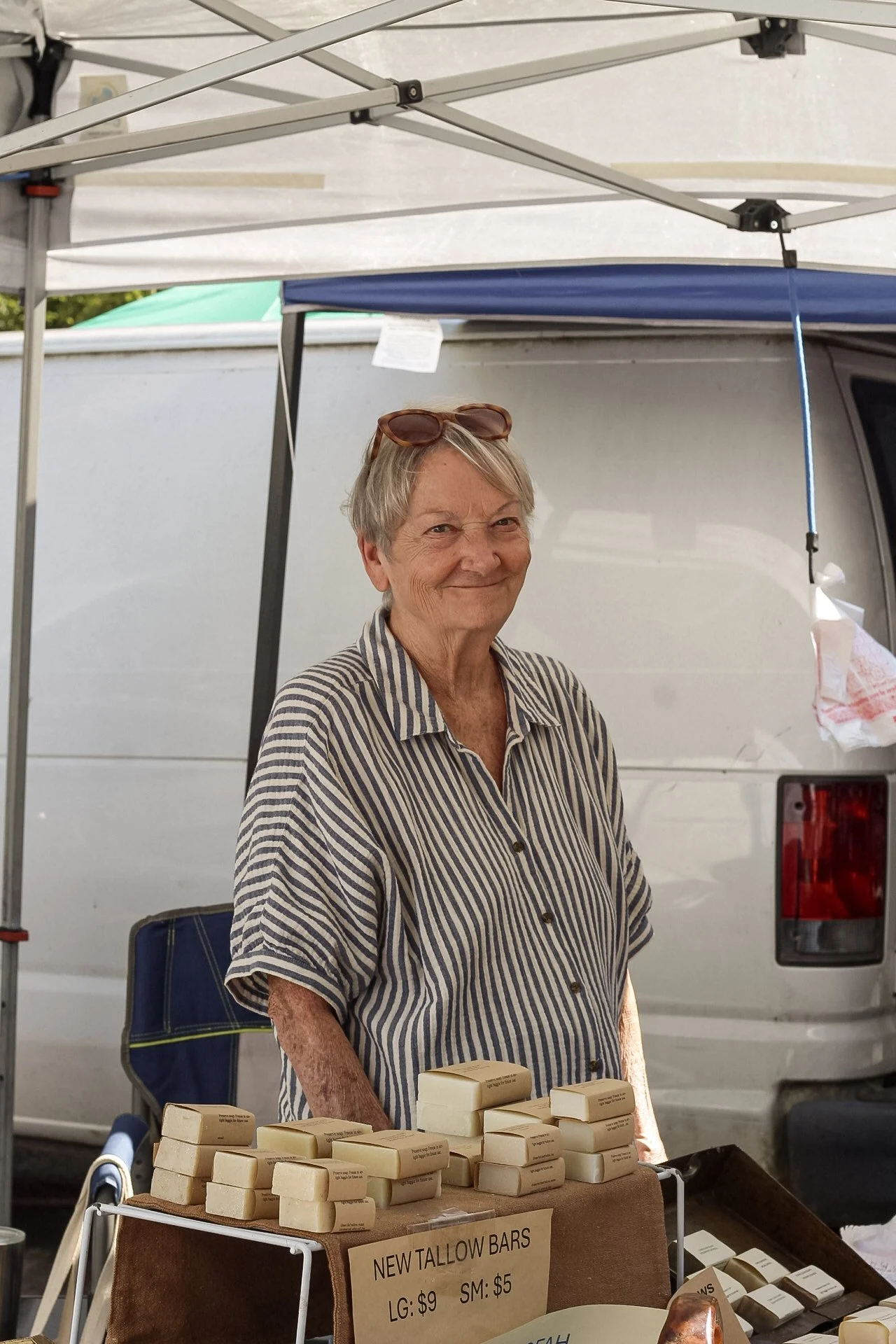 A woman standing behind a table of soap bars at an outdoor market, with a sign reading 'New Tallow Bars' indicating large bars for $9 and small bars for $5, under a tent.