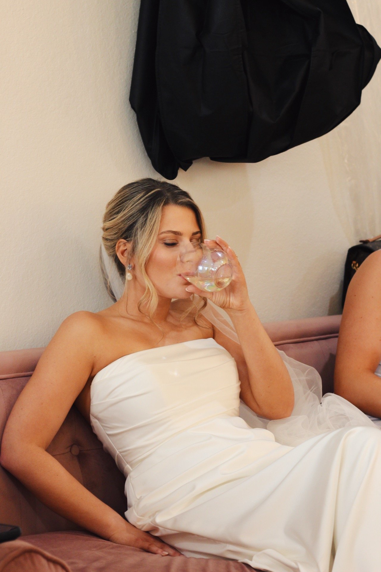 A woman in a white off-shoulder dress sitting on a pink couch, drinking from a glass, with a black bag hanging on the wall behind her.