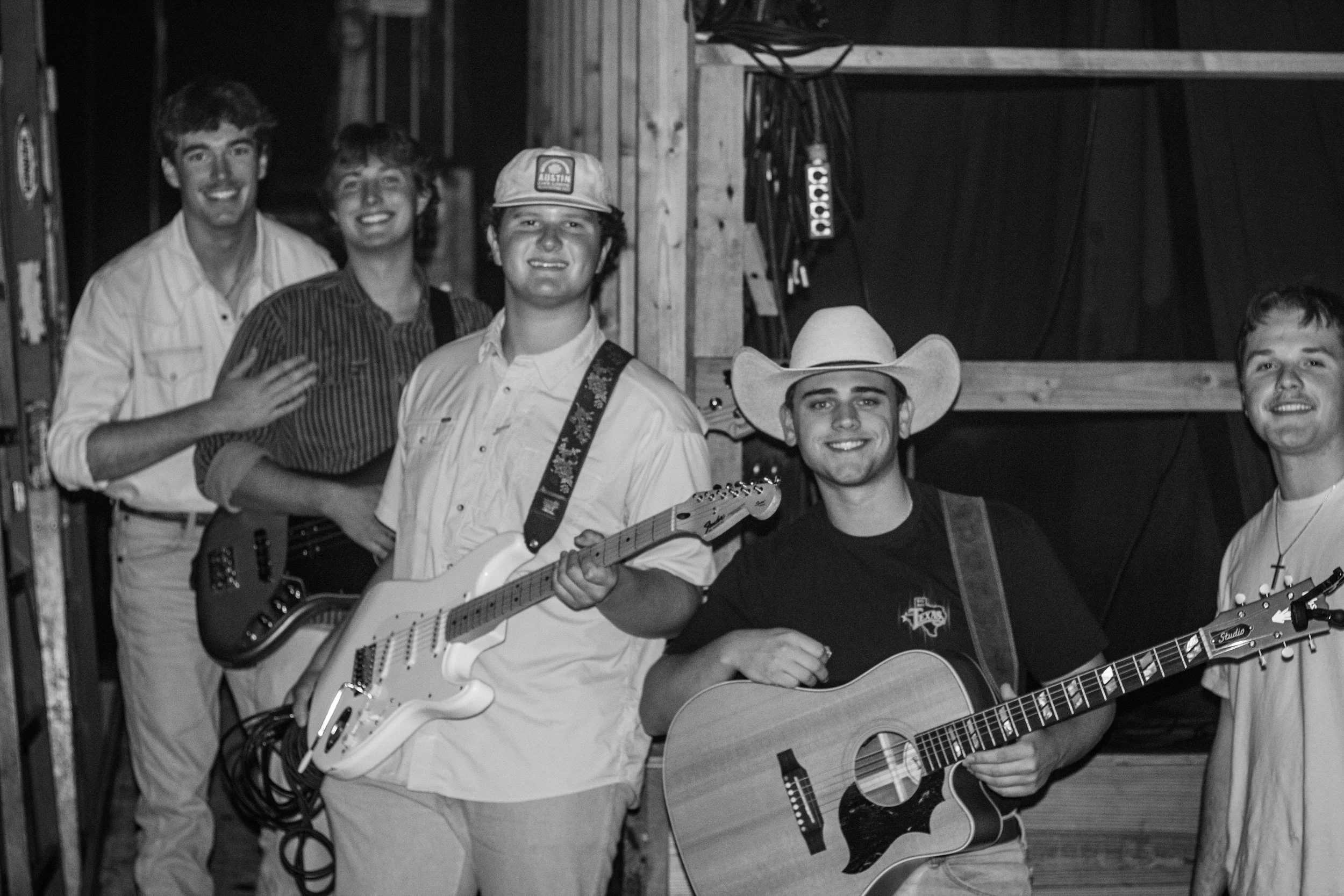 Group of five young men in a rustic setting, some with guitars and one holding an electric guitar, some wearing cowboy hats and casual western-style clothing, smiling and posing for the photo.