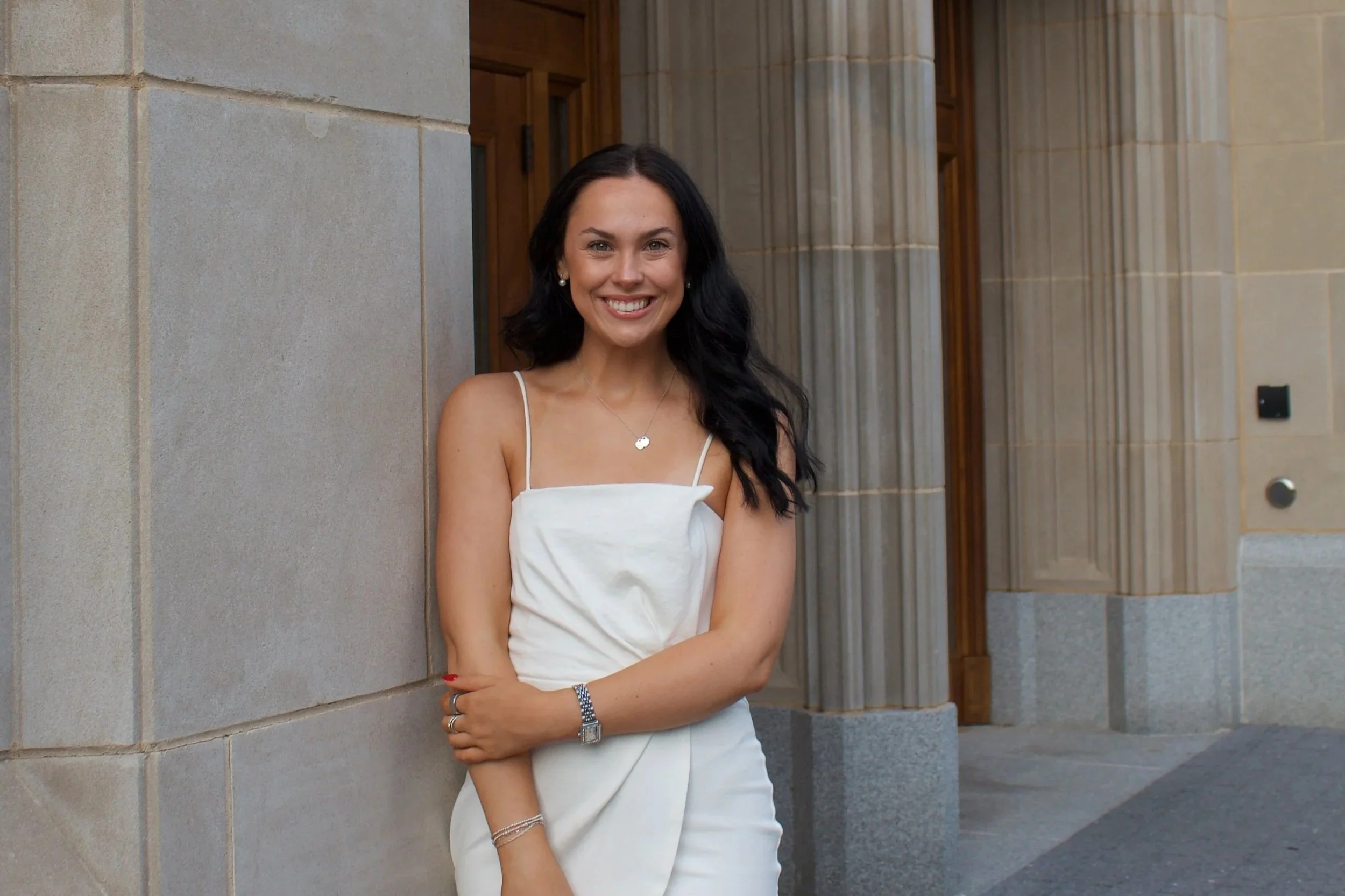 A woman with long black hair wearing a white dress, pearl earrings, and jewelry, smiling and standing outdoors against a stone building with columns.