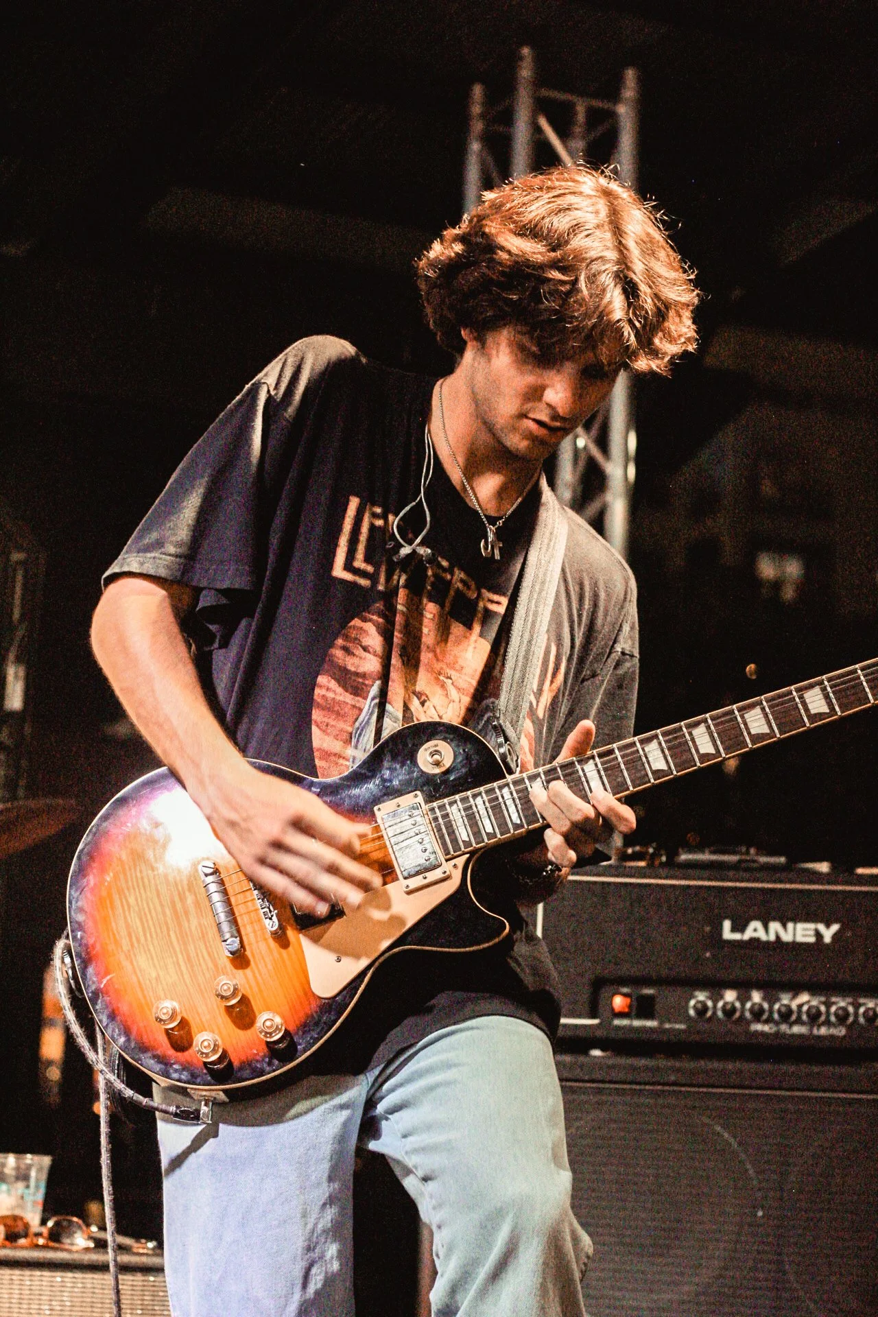 A young man playing an electric guitar on stage, wearing a black band T-shirt, gray pants, a necklace, and earphones, with music equipment in the background.