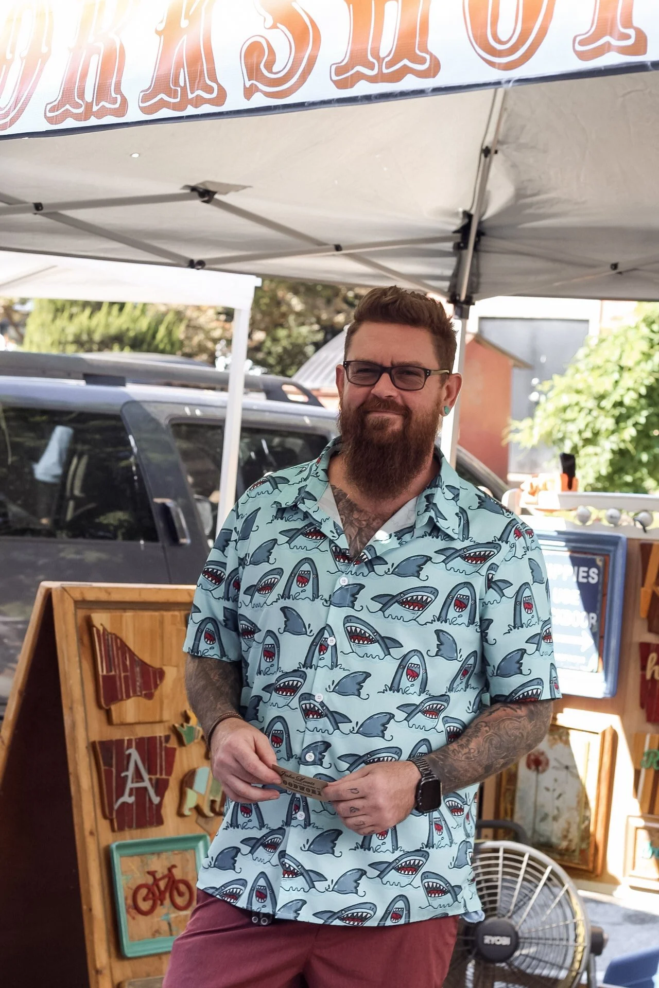 A man with a beard and glasses wearing a light blue Hawaiian shirt with shark patterns stands outdoors under a canopy. He is holding a piece of paper and smiling slightly. Behind him are parked cars, a standing fan, and a sign with painted frames and