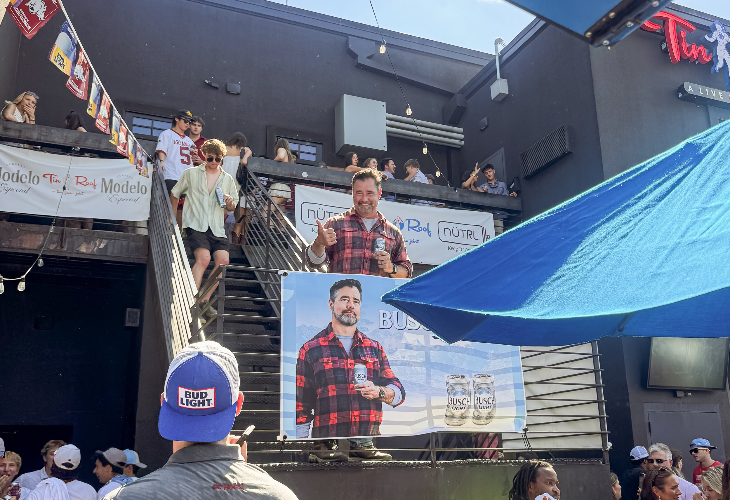 A man in a red plaid shirt holding a Busch Light beer can and giving a thumbs up in front of a large banner with the same man’s image, a crowd of people watching, and a blue Bud Light baseball cap in the foreground. The scene is happening outdoors at