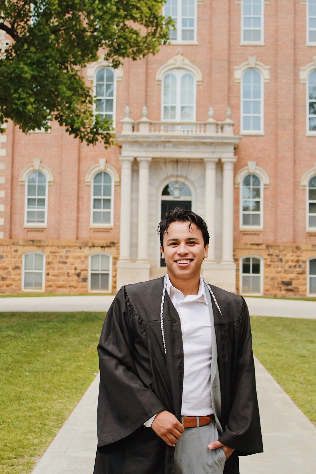 A young man in a graduation gown and cap standing on a sidewalk in front of a historic brick building with large windows and columns, smiling.