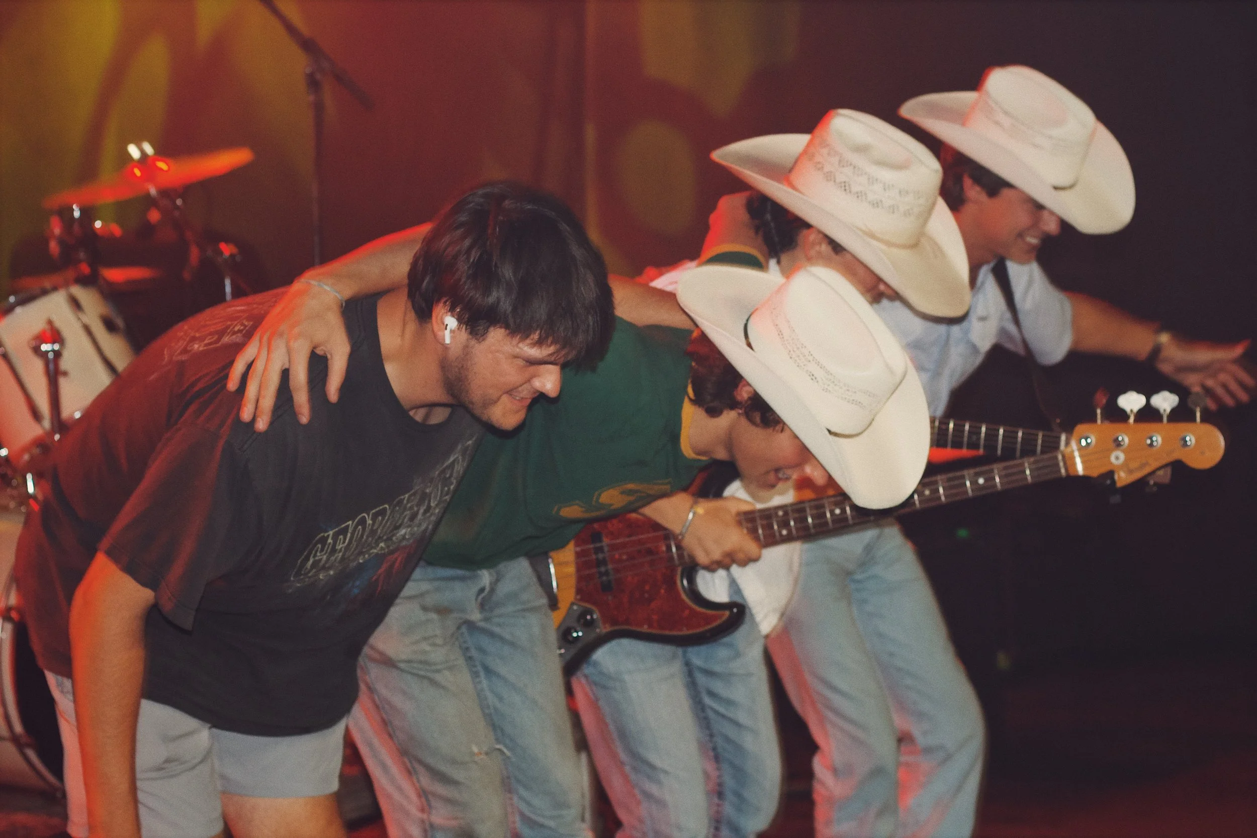 A group of men wearing cowboy hats and casual clothing, standing close together with their arms around each other's shoulders, smiling and bowing their heads in a line.