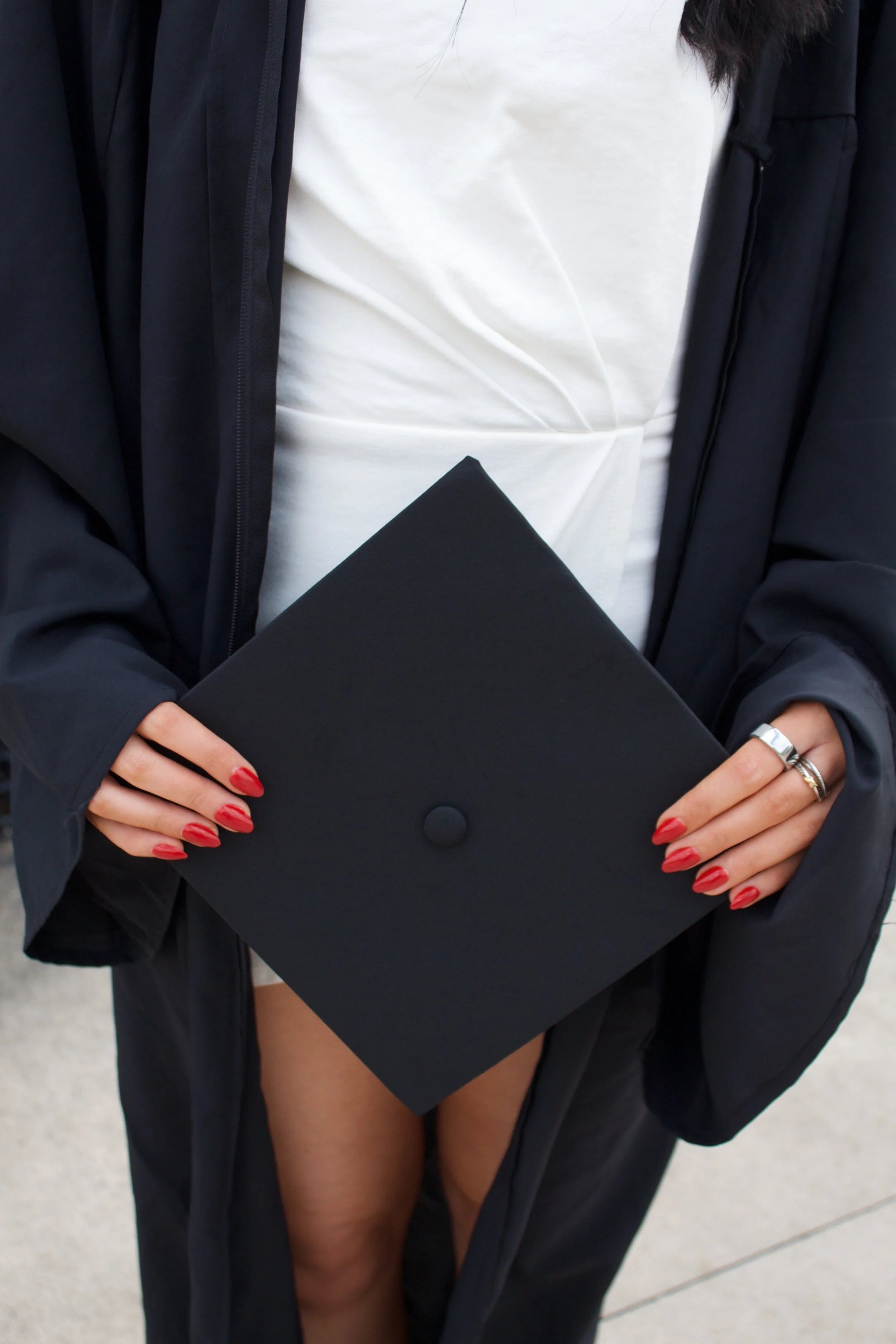 Close-up of a woman holding a black diploma cover, wearing a white gown underneath a black robe, with red painted nails and rings on her fingers.