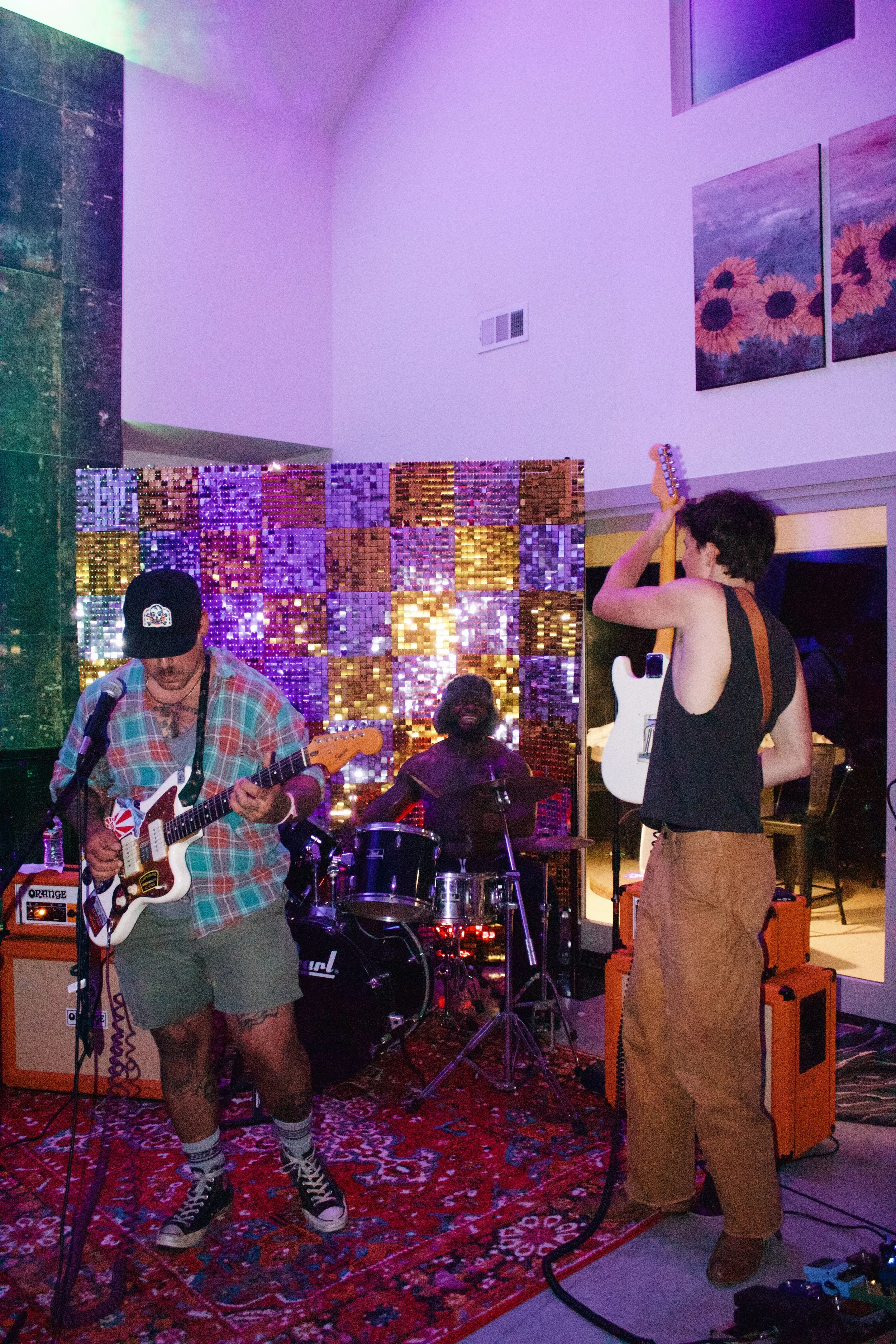 A band of four musicians performing indoors: one playing guitar, one on drums, and two with electric guitars, in a room decorated with paintings and a colorful, shiny partition, with a carpeted floor.