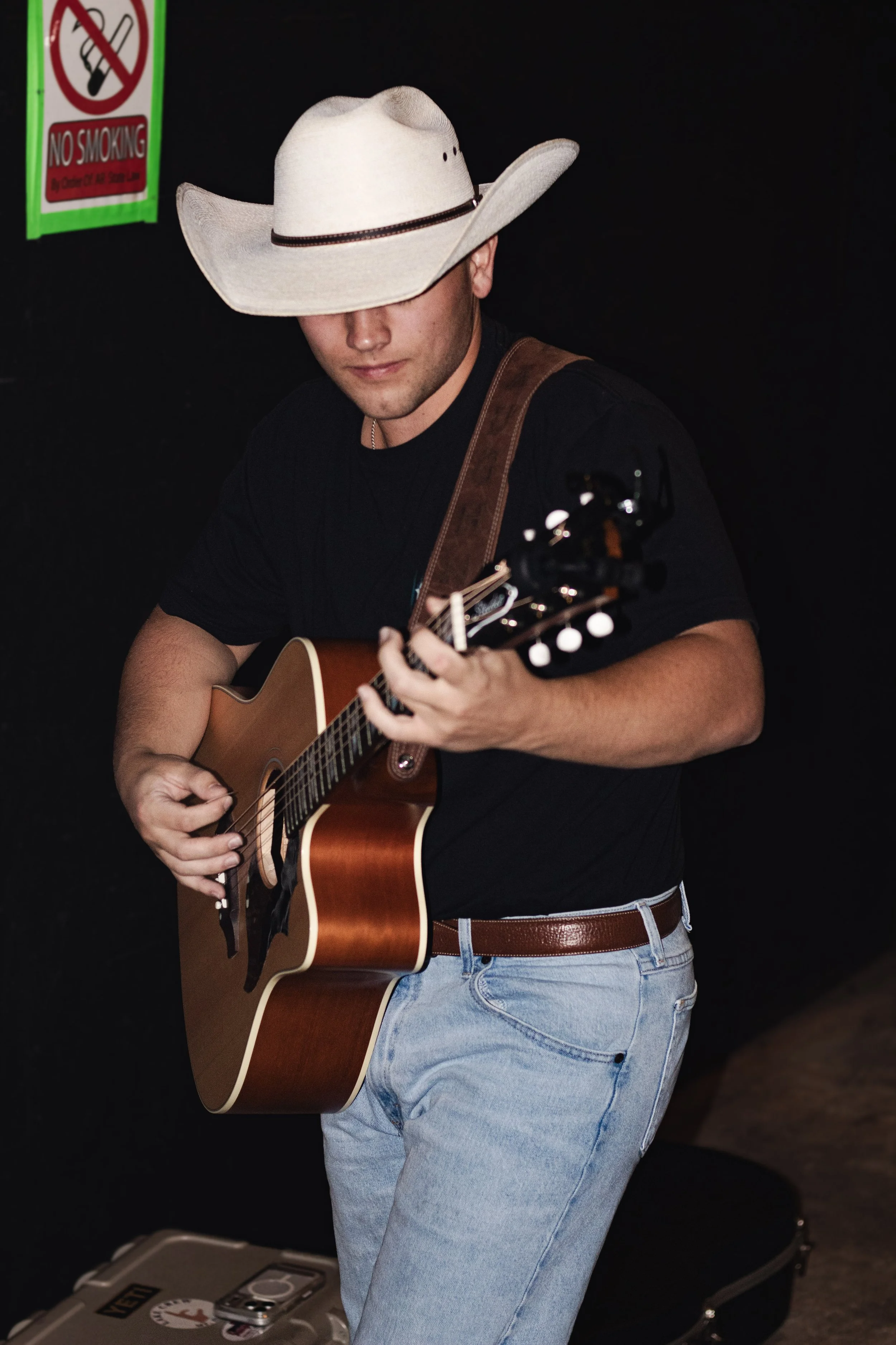 A man wearing a wide-brimmed cowboy hat, black T-shirt, and jeans is playing an acoustic guitar. There is a 'No Smoking' sign in the background and a guitar case nearby.