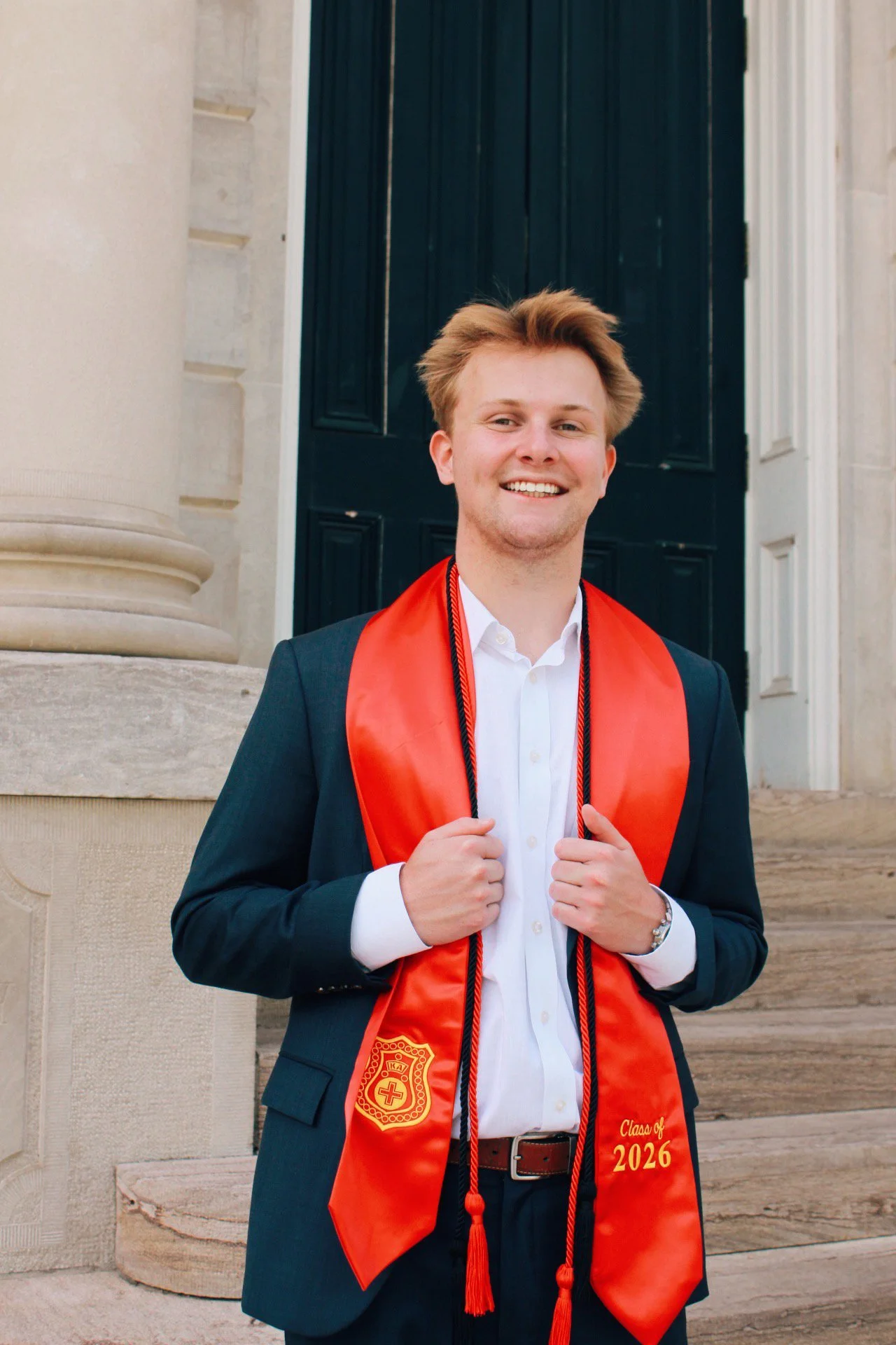 A young man in a suit and white shirt, smiling and holding a red graduation stole with 'Class of 2026' written on it, standing outside a building with stone columns and a dark wooden door.