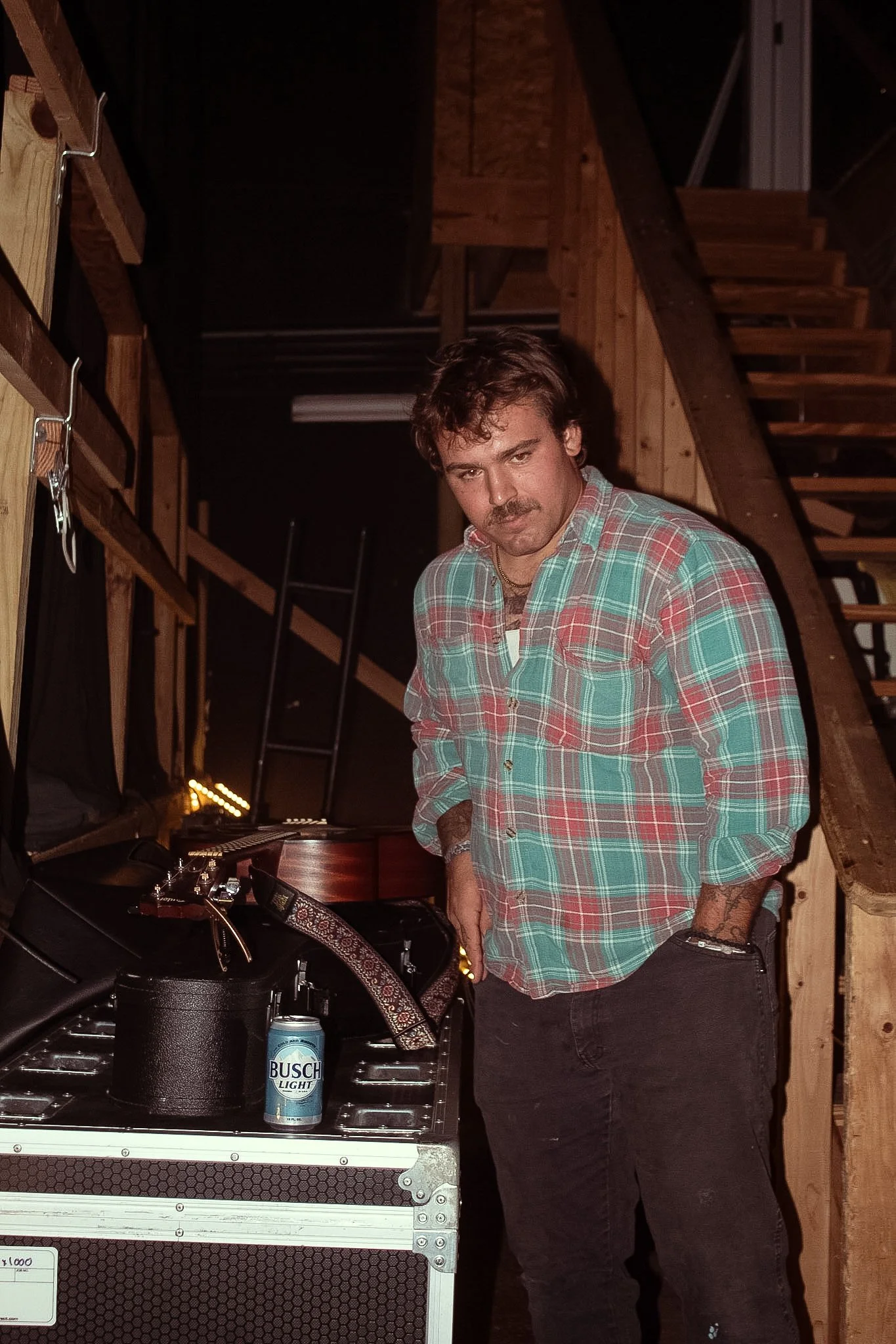 A man with a mustache and curly hair wearing a red and green plaid shirt stands next to DJ equipment in a wooden room. There is a Busch Light beer can on the equipment, and a staircase is visible in the background.