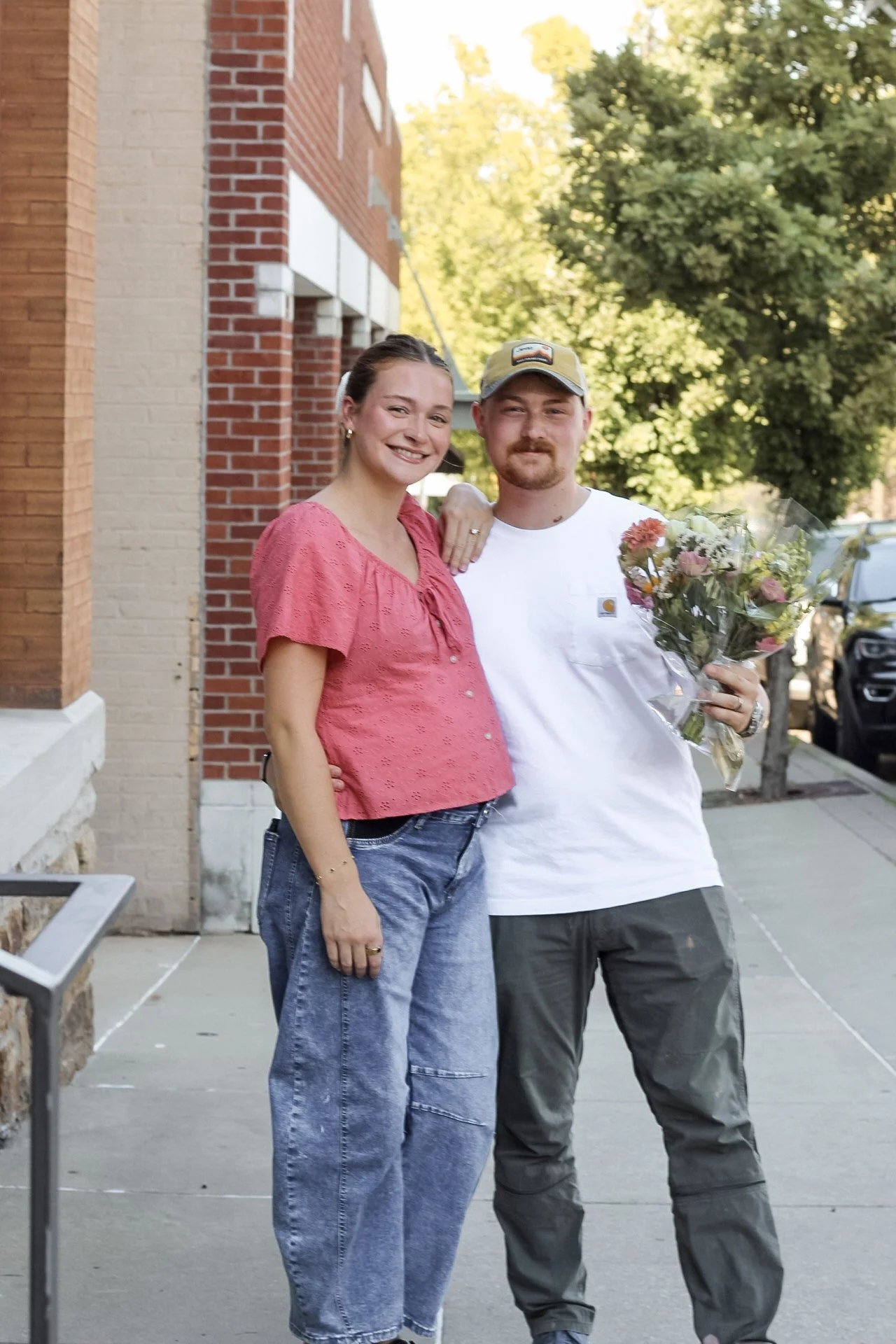 A woman and a man standing outdoors on a sidewalk, smiling at the camera. The woman is wearing a pink blouse and jeans, and the man is wearing a white t-shirt, gray pants, and a cap. The man is holding a bouquet of flowers.