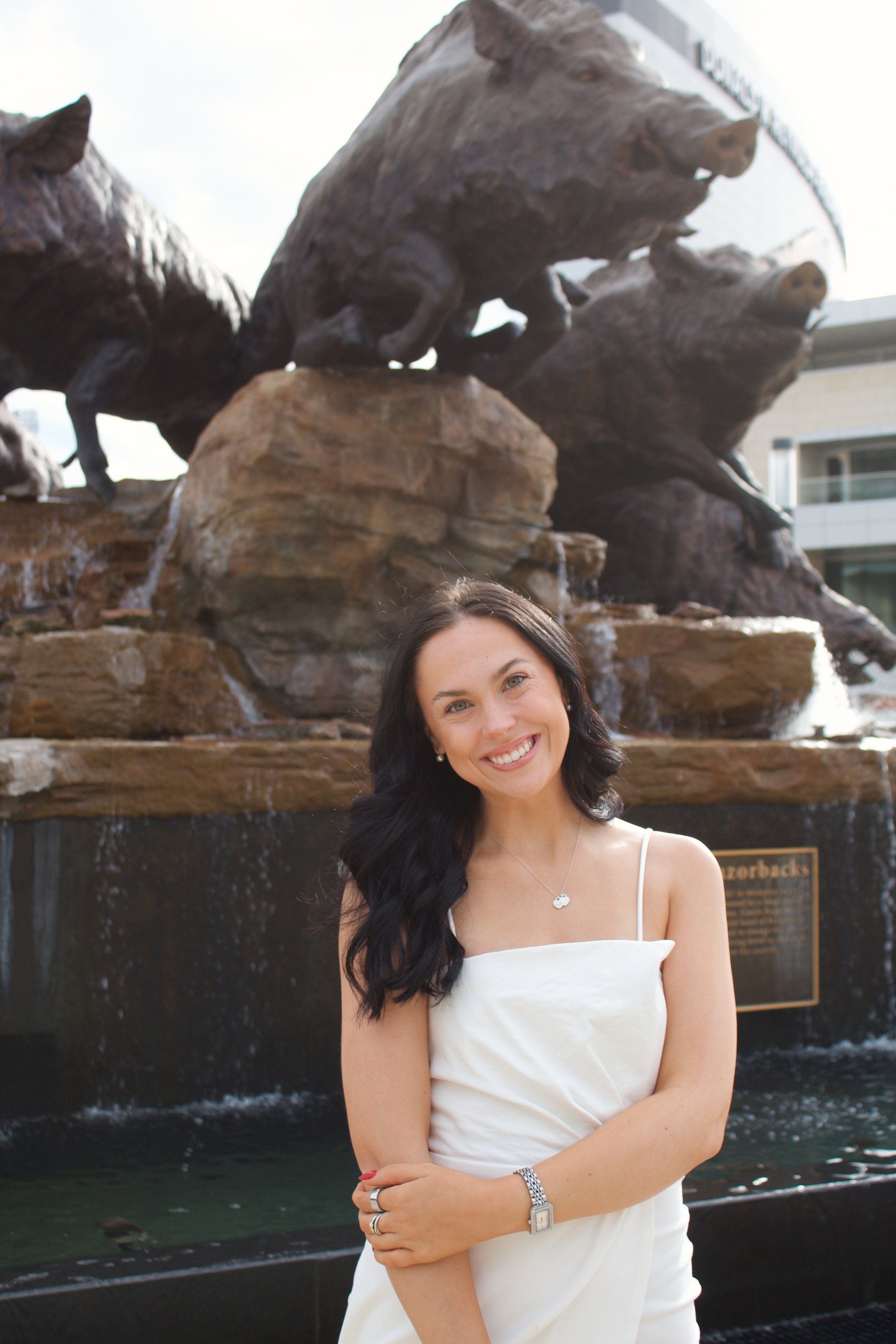 A woman with dark hair wearing a white dress stands in front of a fountain with a bronze sculpture of wild boars on rocks, in an urban setting.