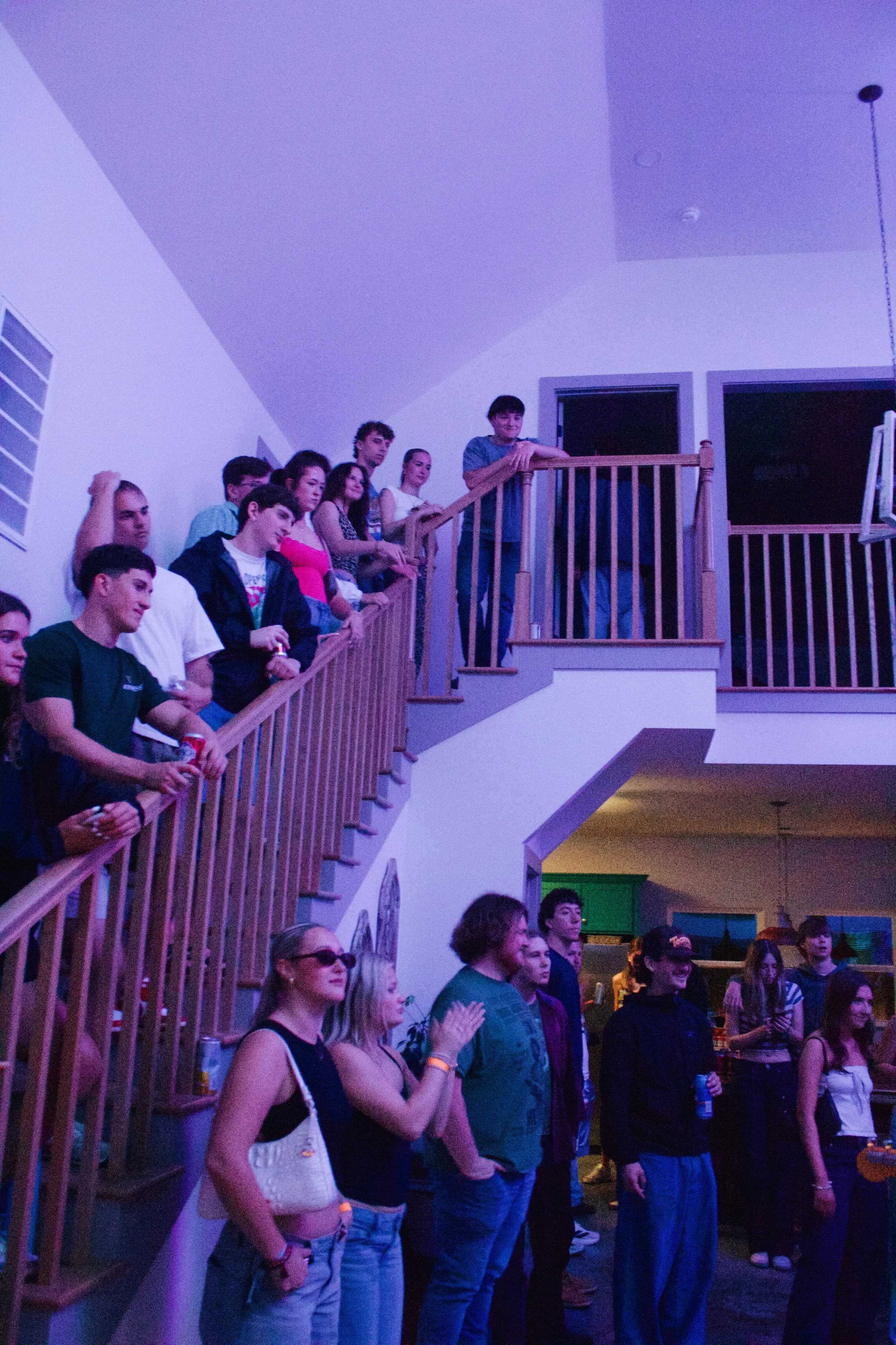 Group of young people watching an event at an indoor venue, standing along a wooden railing, some clapping, with a colorful background and a man leaning on the railing at the top of the stairs.