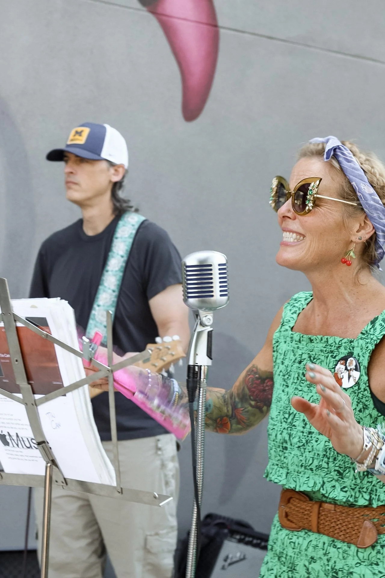 A woman with blonde curly hair, wearing sunglasses, a blue bandana, earrings shaped like cherries, and a green dress, appears to be singing or speaking into a vintage microphone. A man next to her is playing a guitar, wearing a black t-shirt, a white