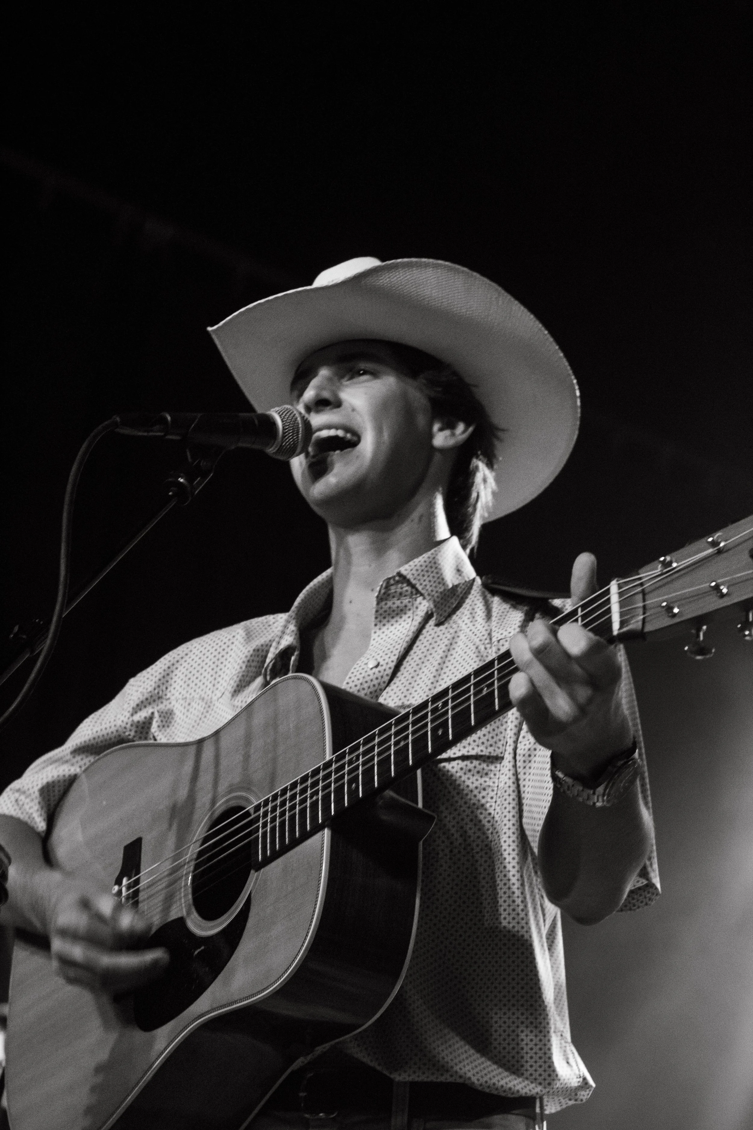 A man wearing a cowboy hat singing into a microphone and playing an acoustic guitar on stage.