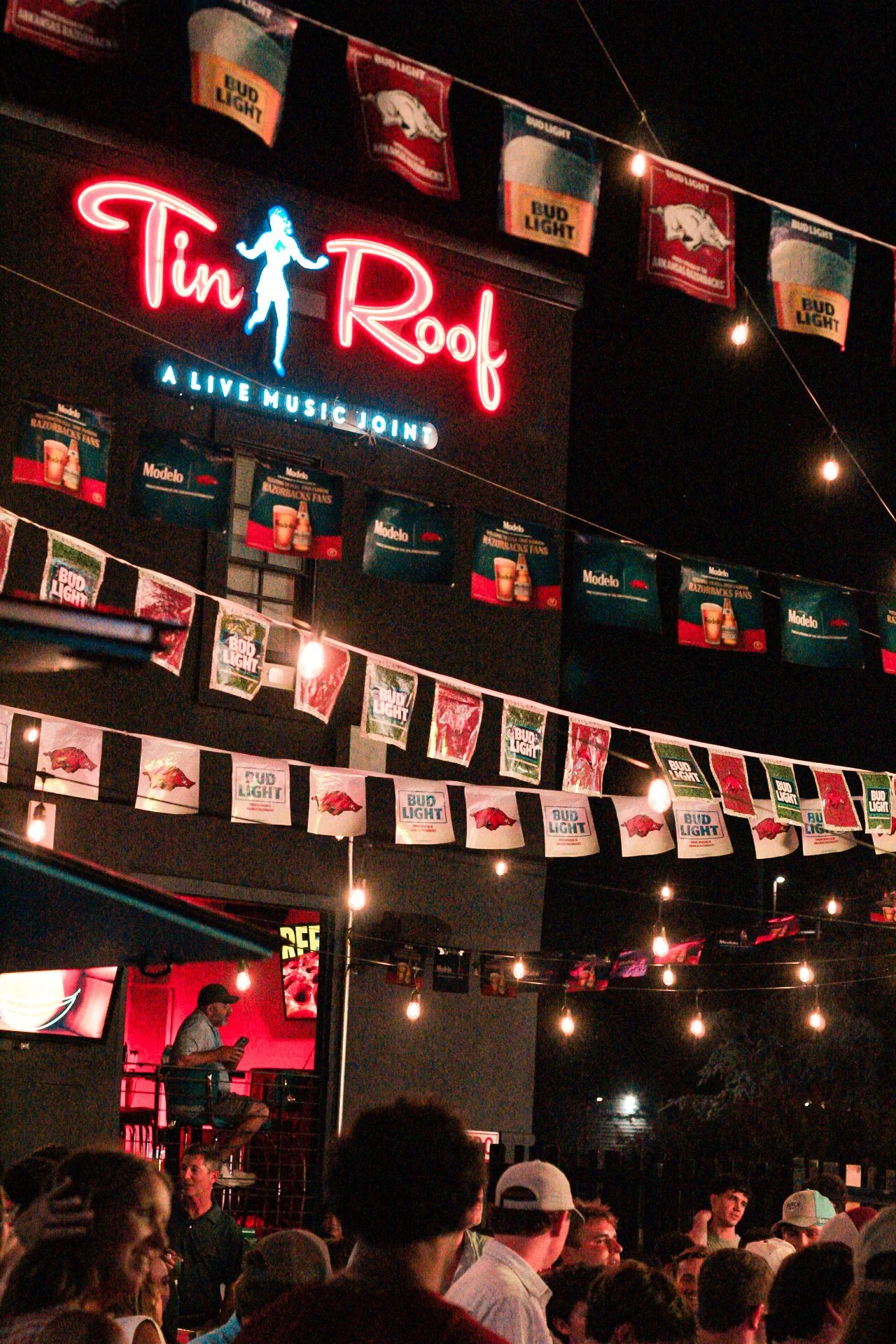 Night scene at a live music venue called Tin Roof with a crowd of people, string lights, and patriotic banners featuring Bud Light and Arkansas Razorbacks.