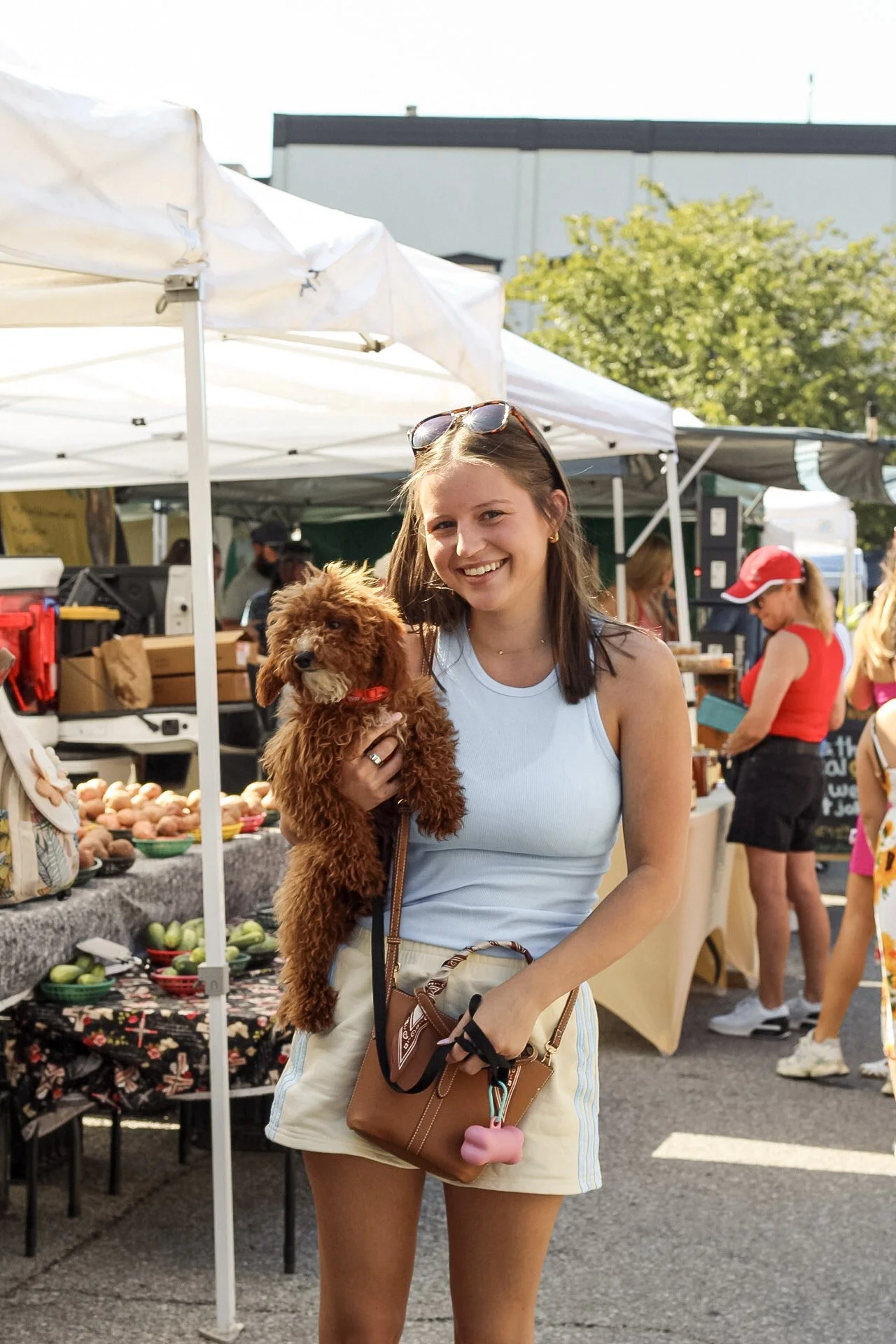 A young woman holding a small brown curly-haired dog at an outdoor market, smiling at the camera, with stalls and people in the background.